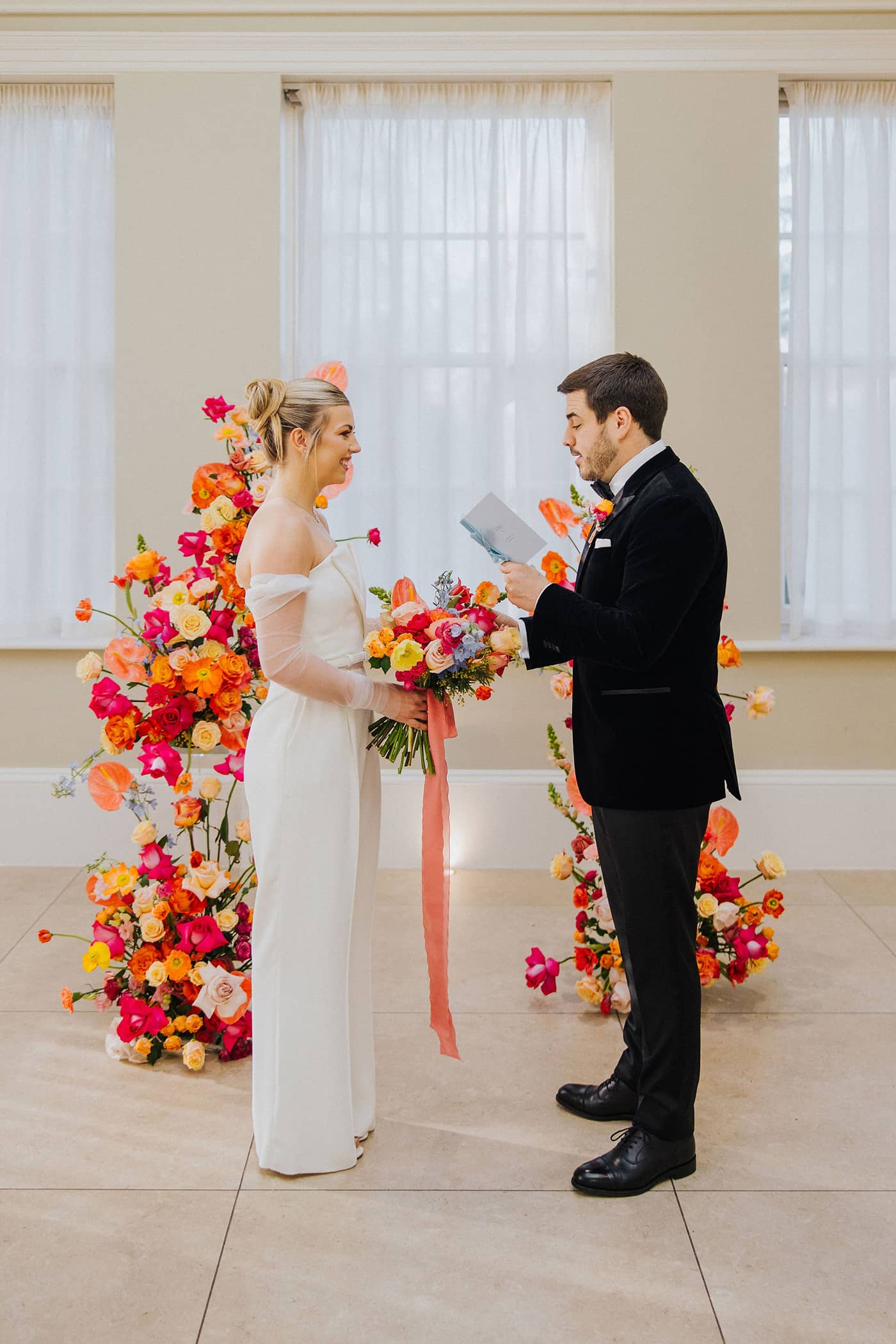 A bride dressed in a white jumpsuit and a groom in a black tuxedo exchanging vows in front of a bright and vibrant floral arch at Saltmarshe Hall. The bride is carrying a bright and colourful wedding bouquet and the groom is holding a pastel blue vow book.