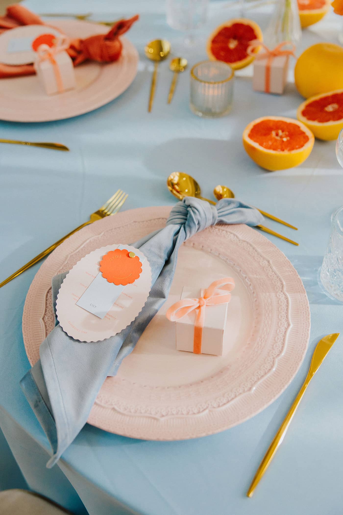 A wedding table setting with pastel pink charger plates, pastel pink favour boxes with a coral velvet ribbon and a blue. napkin, all sitting on a pastel blue tablecloth with gold cutlery.