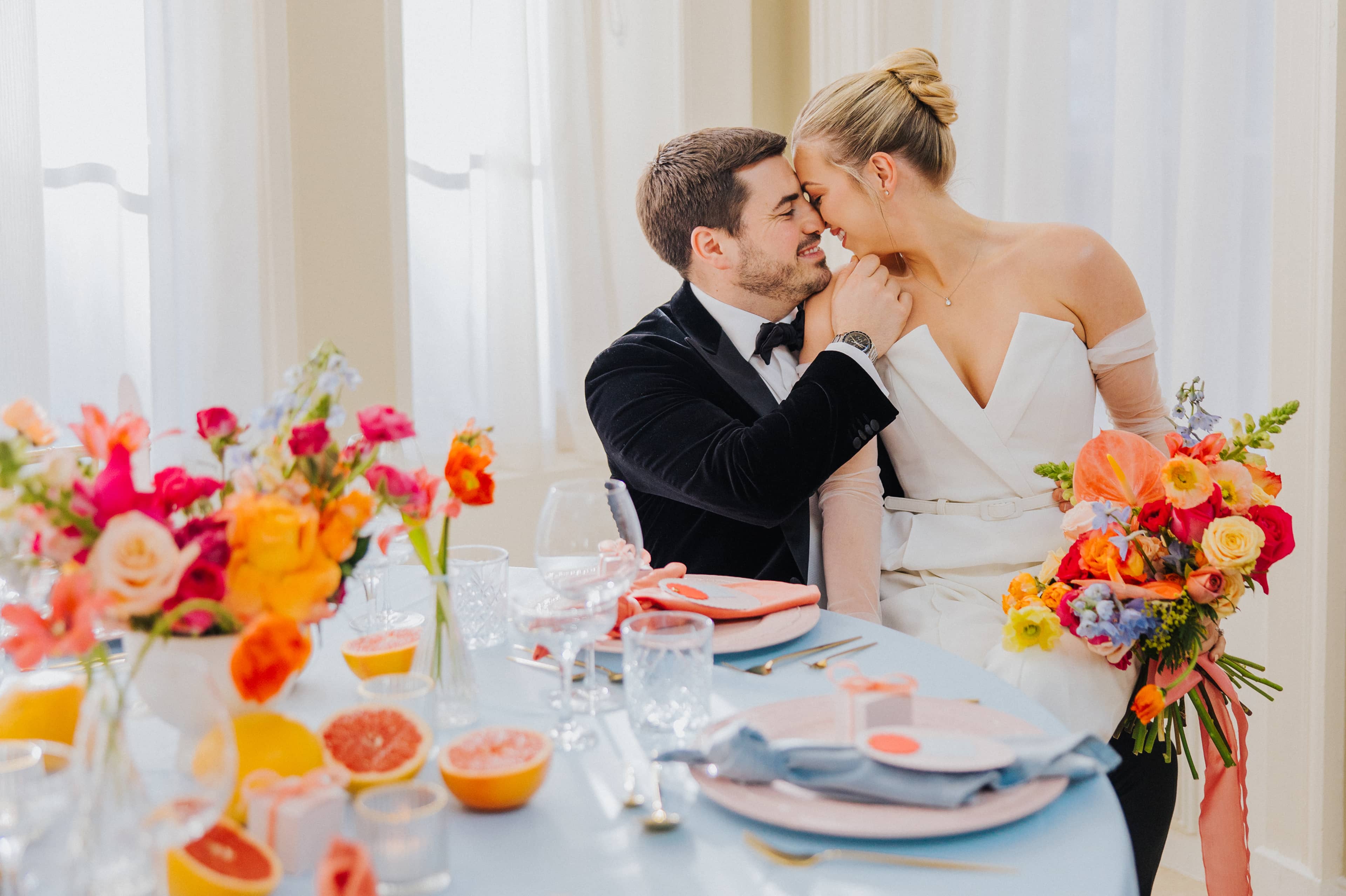A bride dressed in a white jumpsuit and a groom in a black tuxedo sitting at a round wedding table at Saltmarshe Hall, embracing each other. The bride is carrying a bright and colourful wedding bouquet.