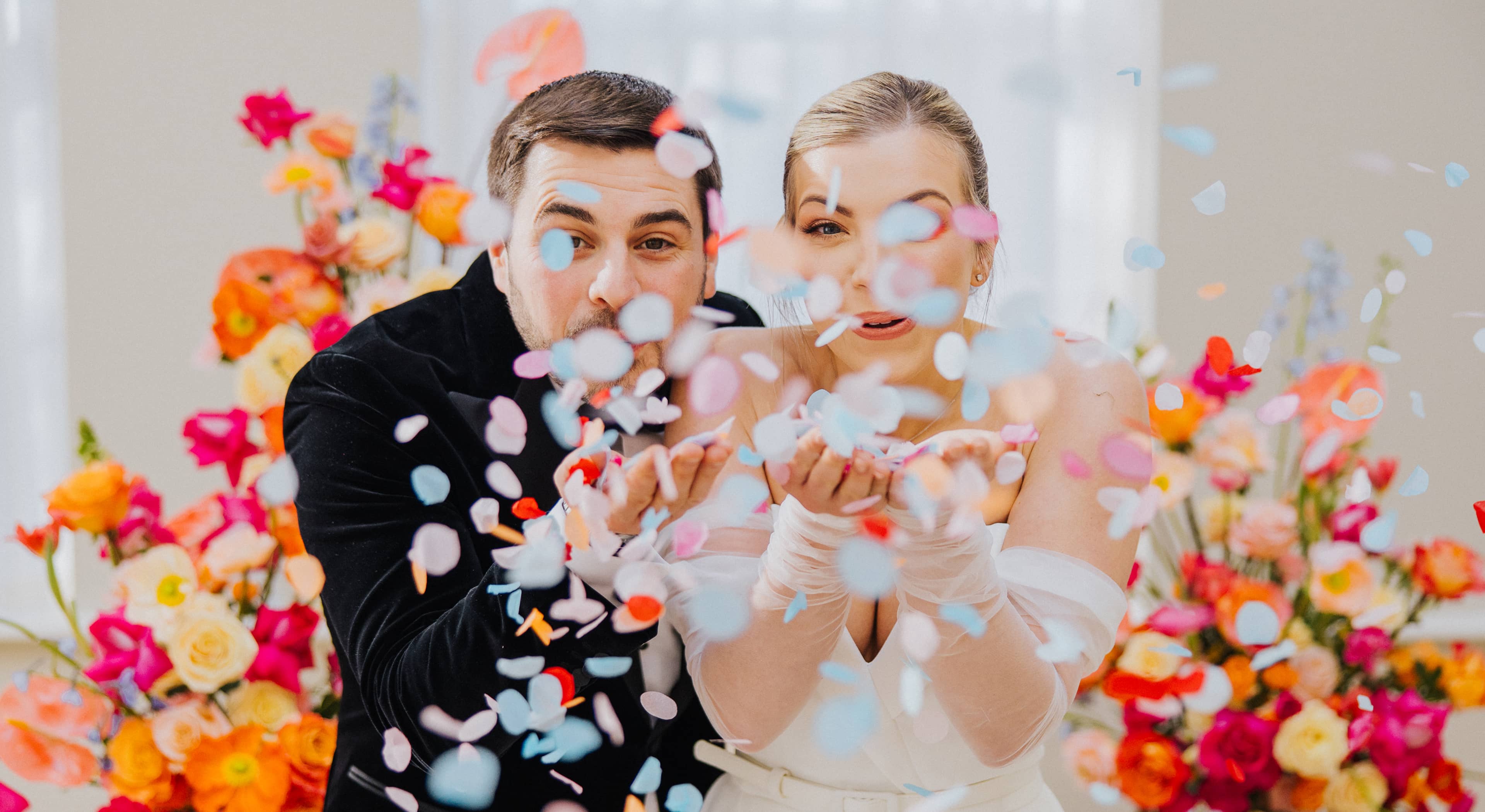 A bride and groom blowing pastel pink, blue and orange confetti out of their hands in front of a colourful flower arch at Saltmarshe Hall.