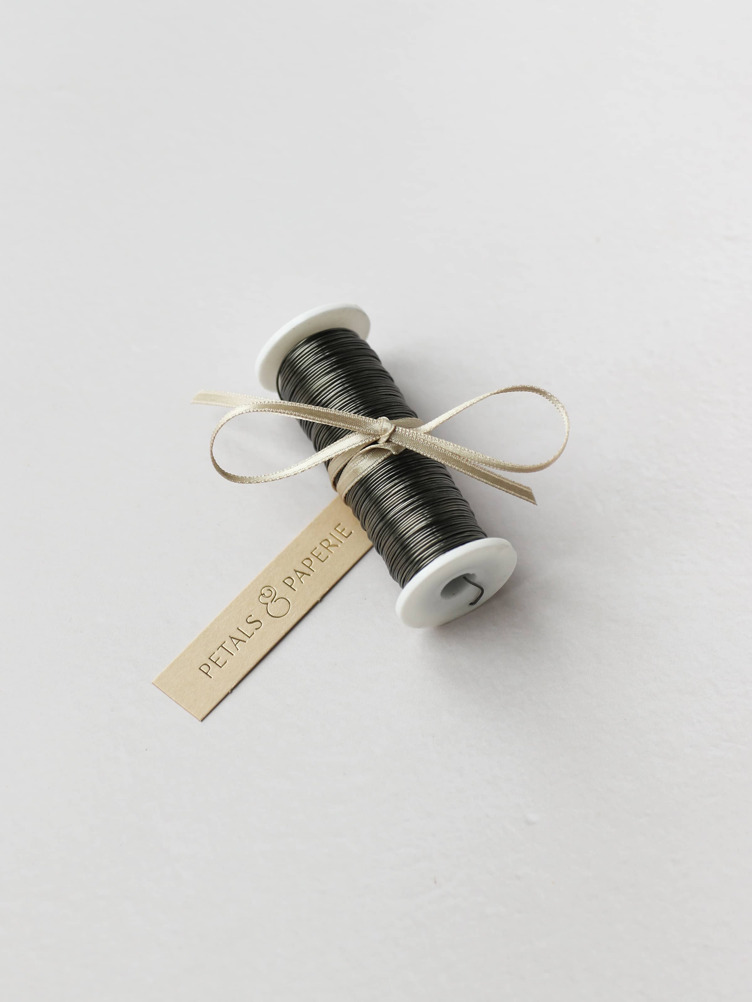 A spool of silver reel wire for Christmas wreath making, tied with a cream satin ribbon and Petals & Paperie gift tag on a neutral background.