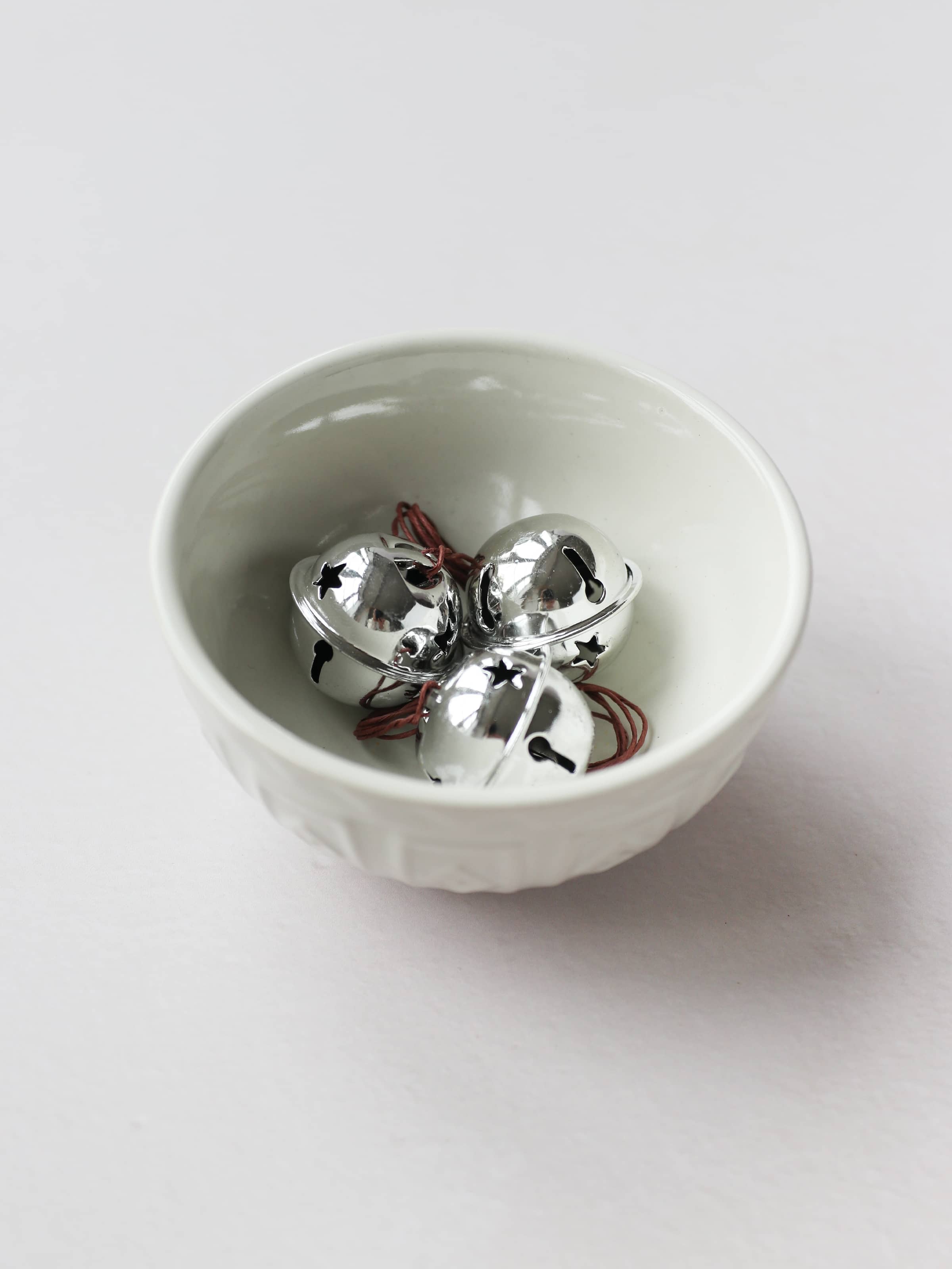 A small cream bowl with 3 silver jingle bells, sitting on a light grey background