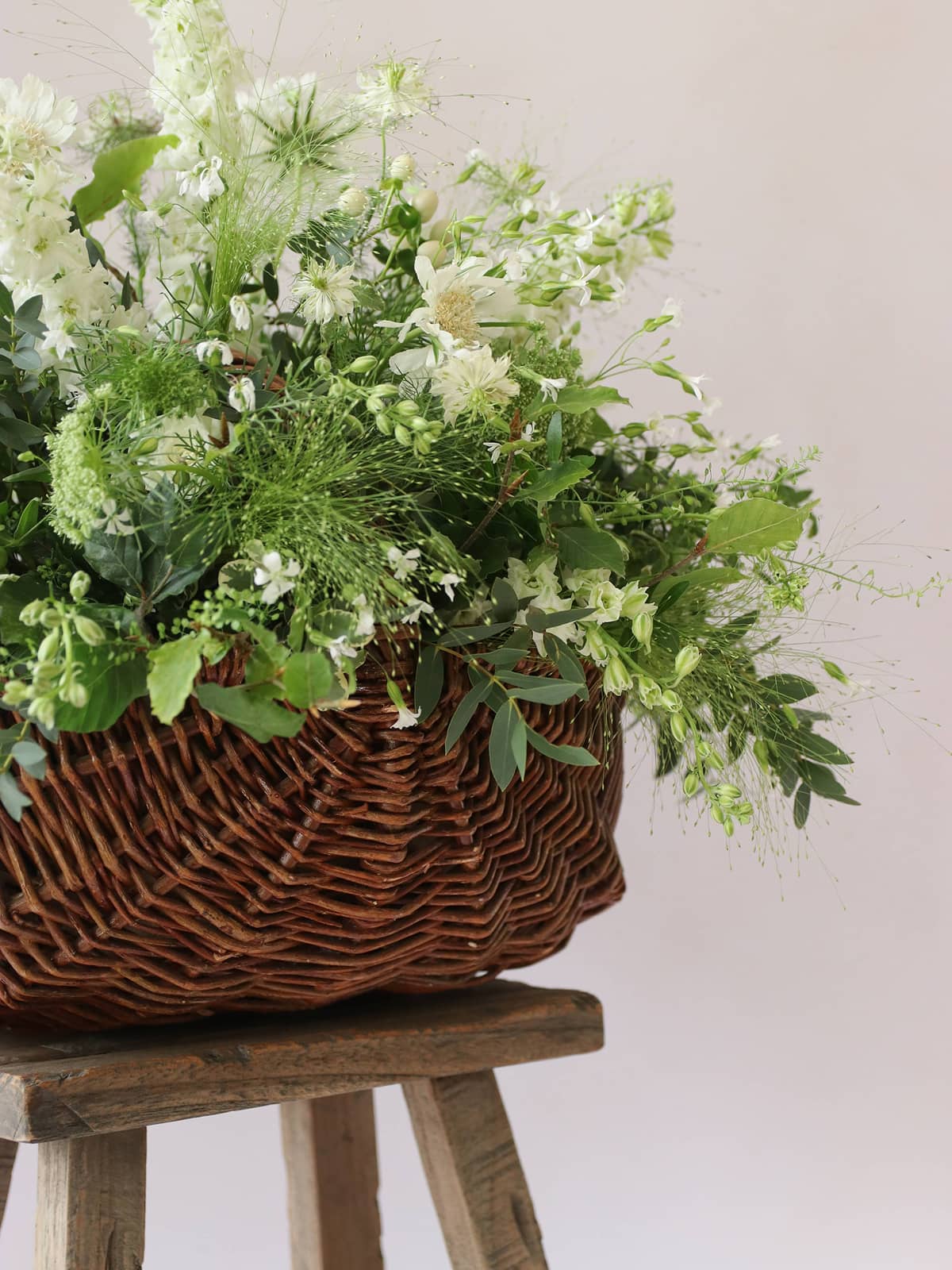 Wicker basket filled with greenery and white flowers on a wooden stool against a light background