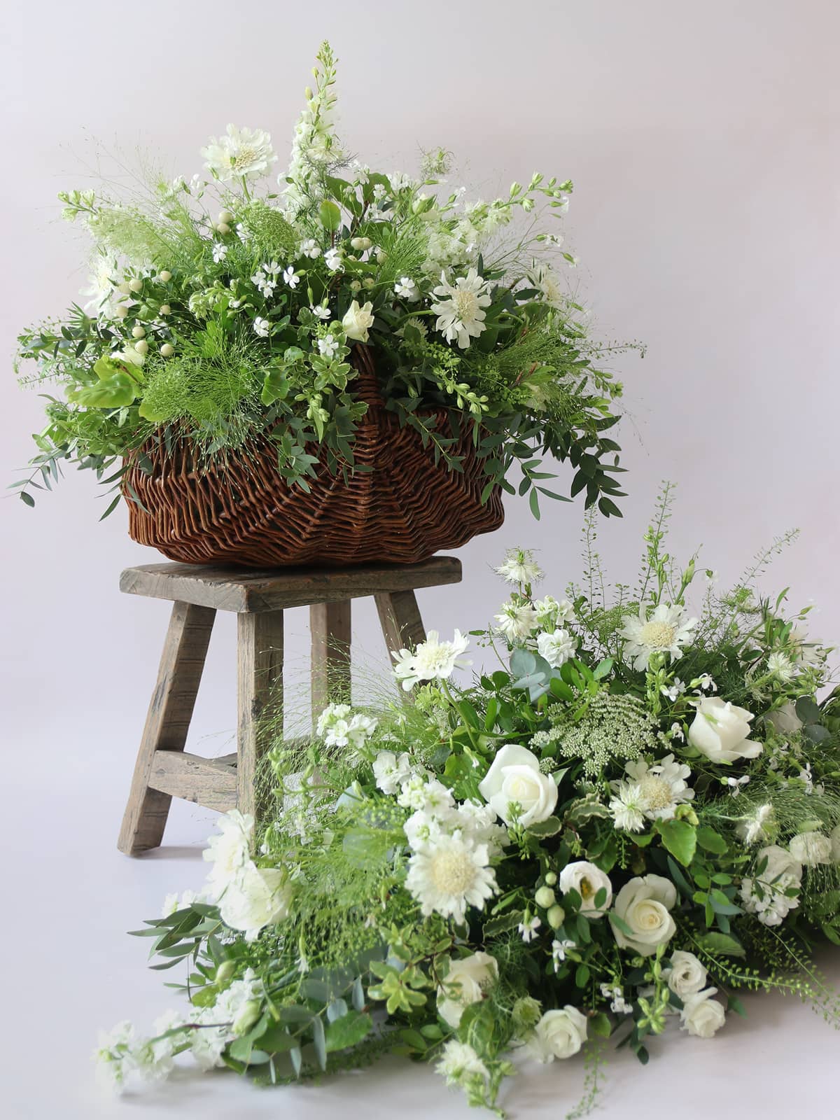 Wicker basket of flowers on a wooden stool with additional double ended coffin spray arrangement on a white background