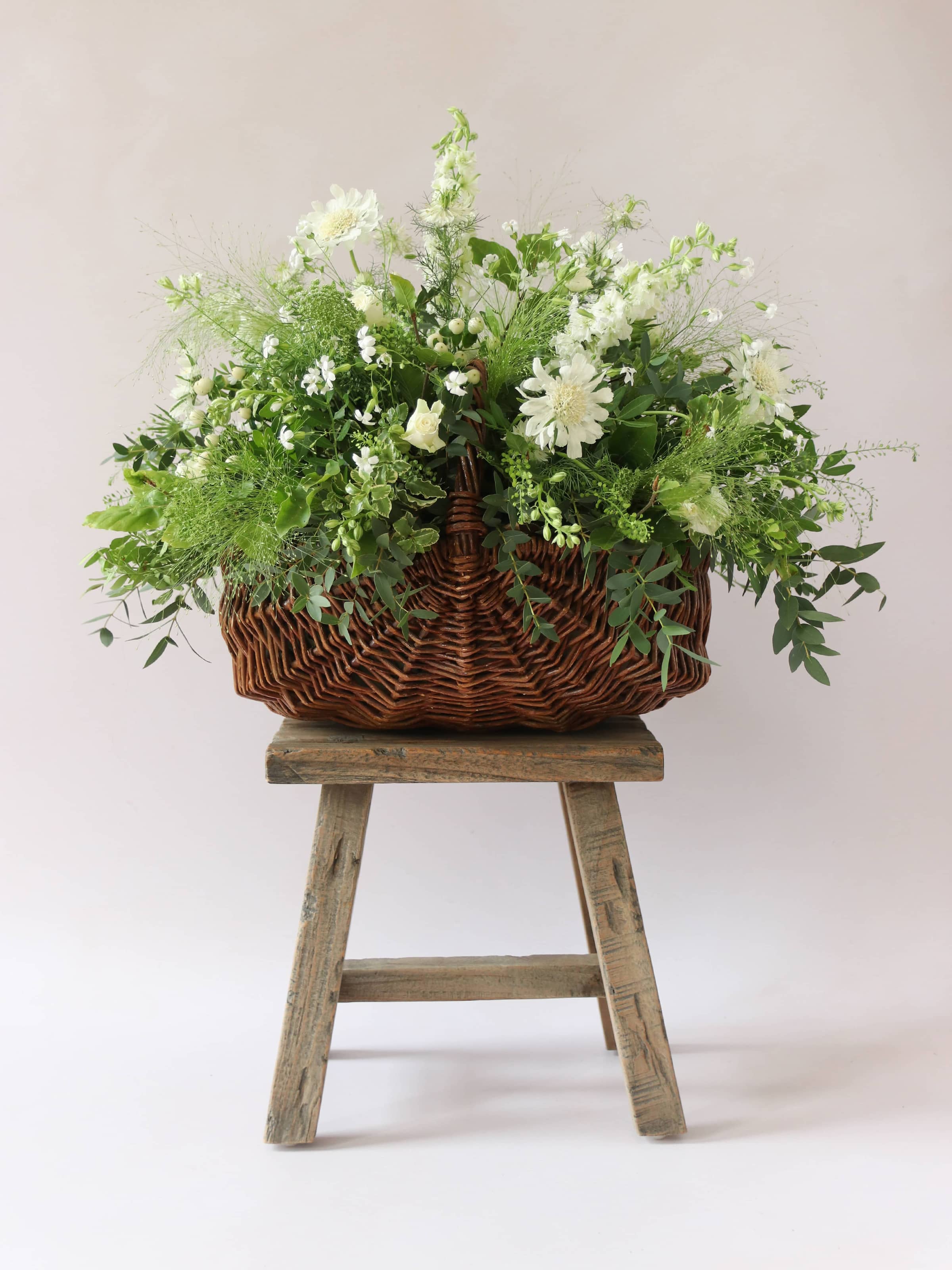 A wicker basket arrangement featuring white and green flowers and foliage. The basket arrangement is sitting on a rustic wooden stool against a neutral background and features white Roses, Ammi, Stocks and Lisianthus.