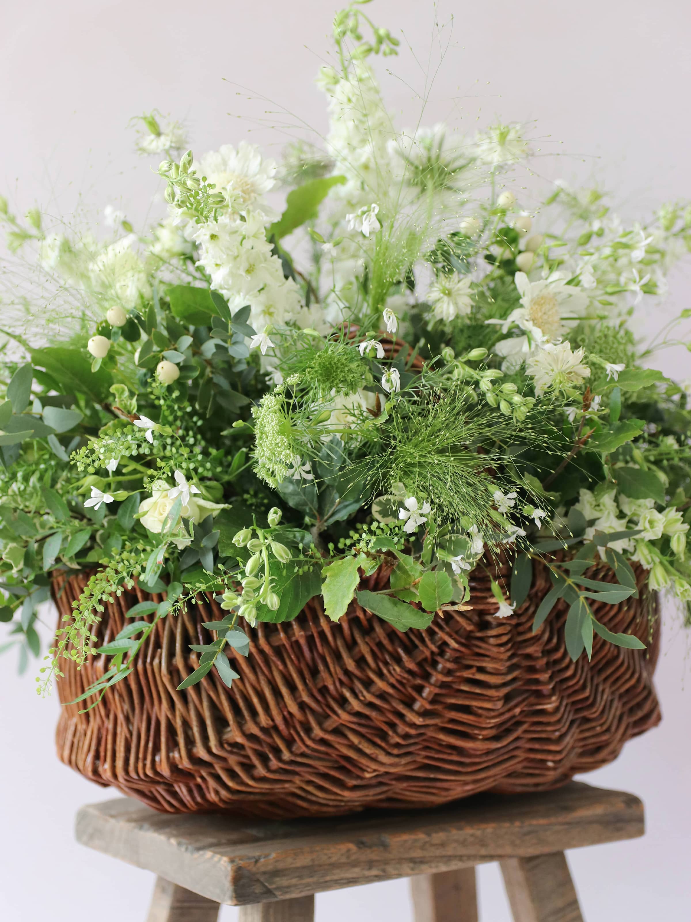 A side view of a wicker basket arrangement featuring white and green flowers and foliage. The basket arrangement is sitting on a rustic wooden stool against a neutral background and features white Roses, Ammi, Stocks and Lisianthus.