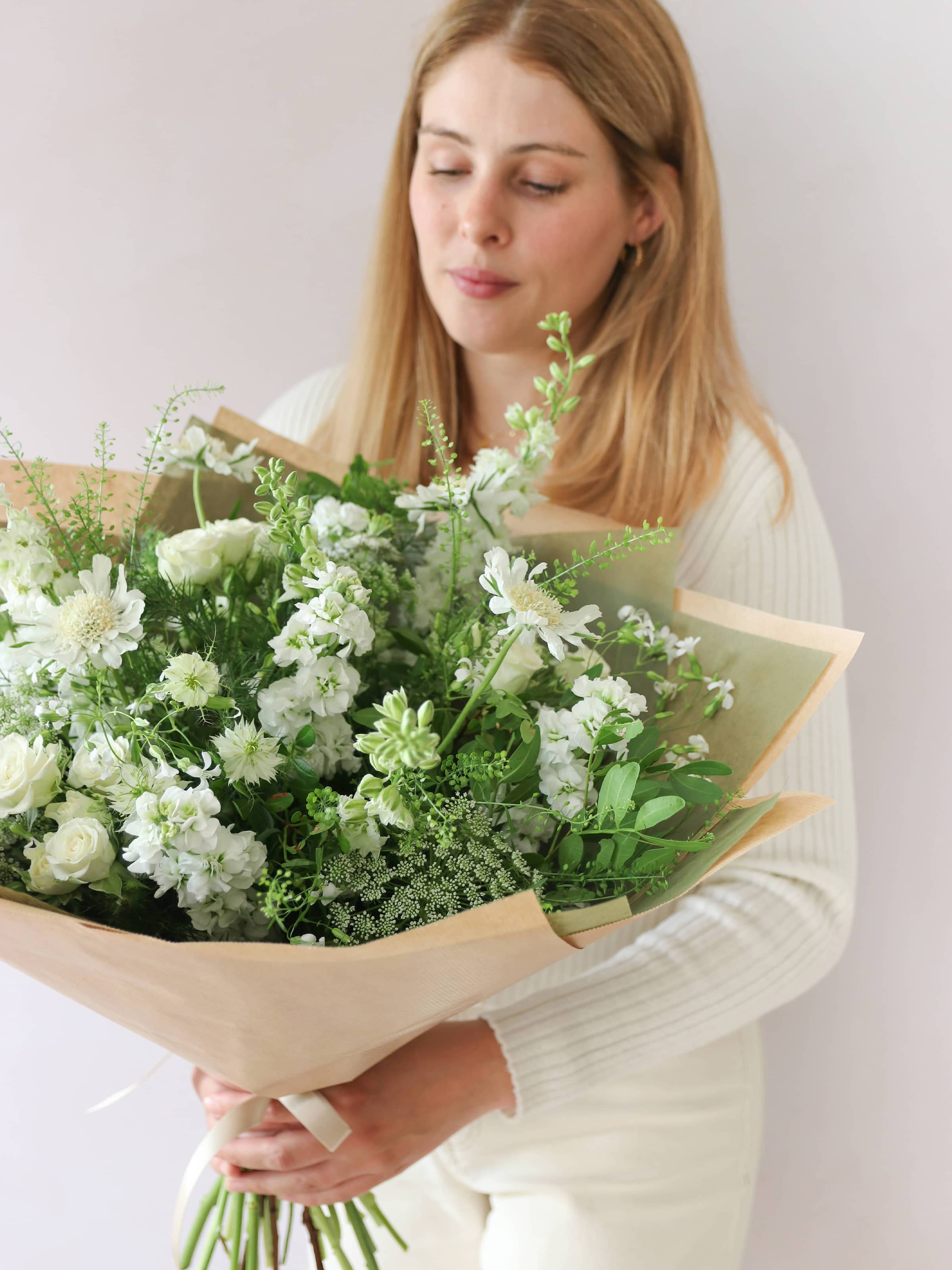 A woman wearing a white jumper and cream jeans, looking down at a white and green gift-wrapped bouquet of flowers including Delphiniums, Stocks, Roses and Ammi.