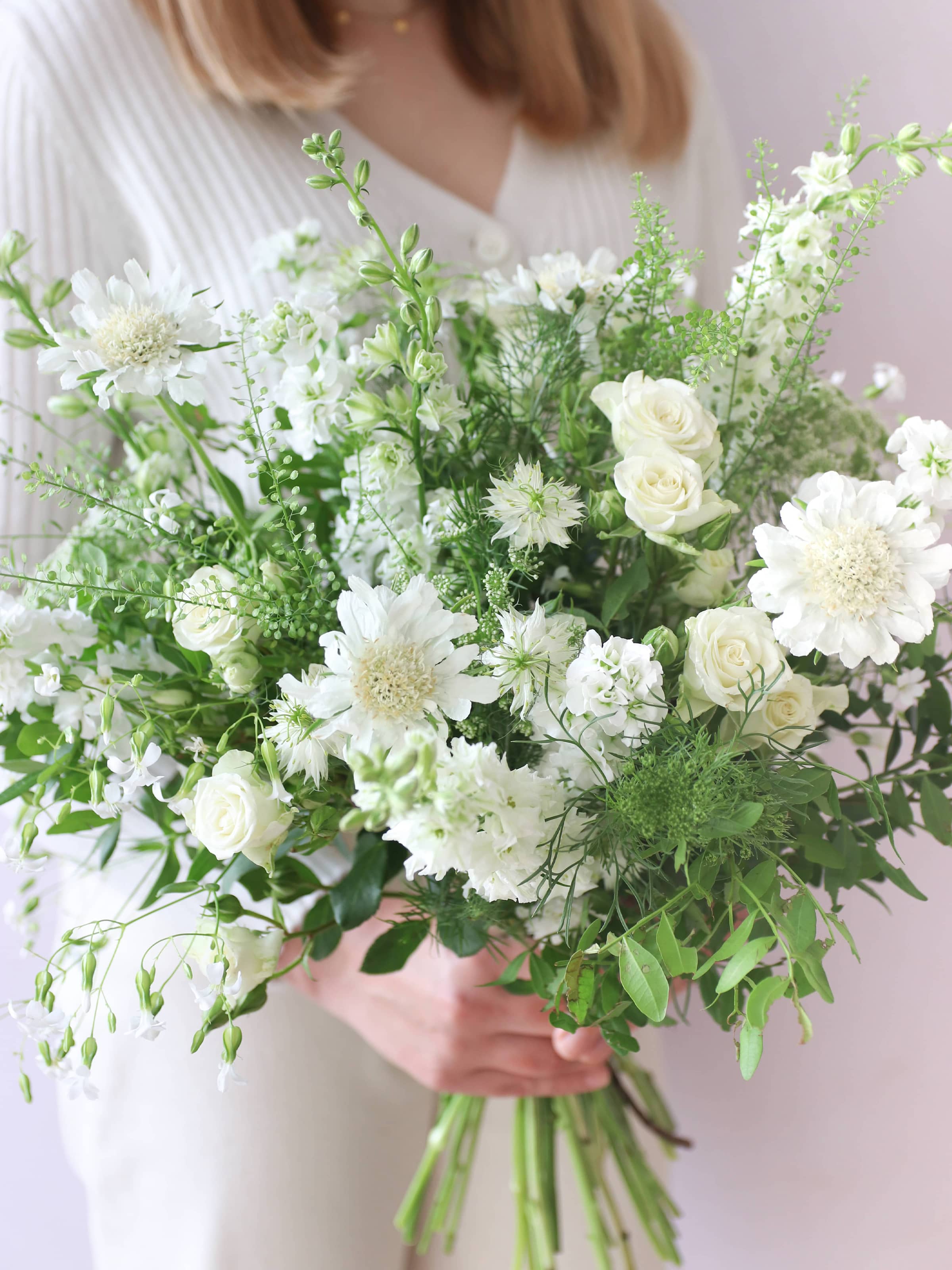 A woman wearing a white jumper and cream jeans, holding a white and green bouquet of flowers including Delphiniums, Stocks, Roses and Ammi in both hands against a neutral background.