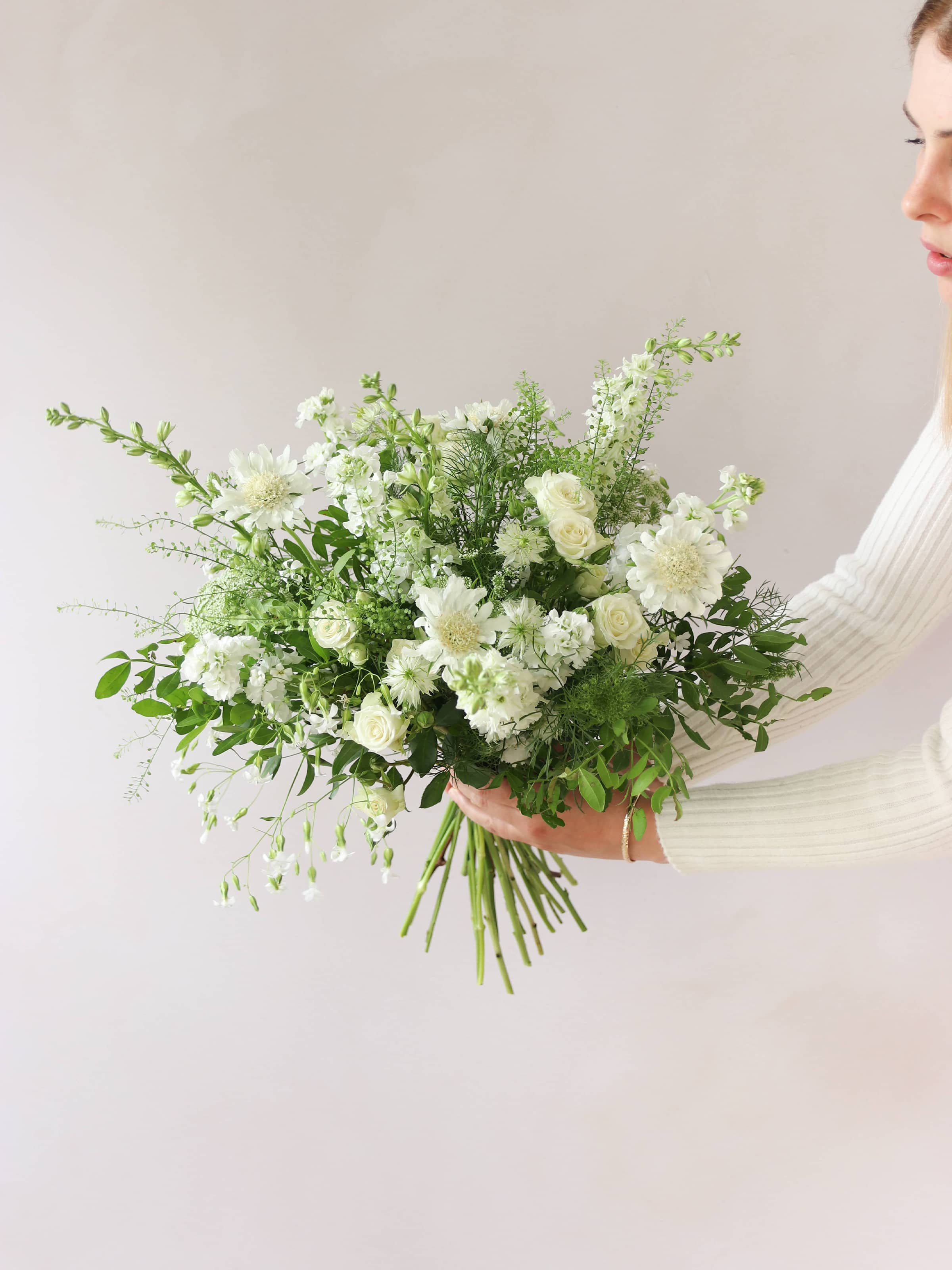 A woman holding a white and green bouquet of flowers out to the side against a neutral background. The bouquet includes Delphiniums, Stocks, Roses and Ammi.