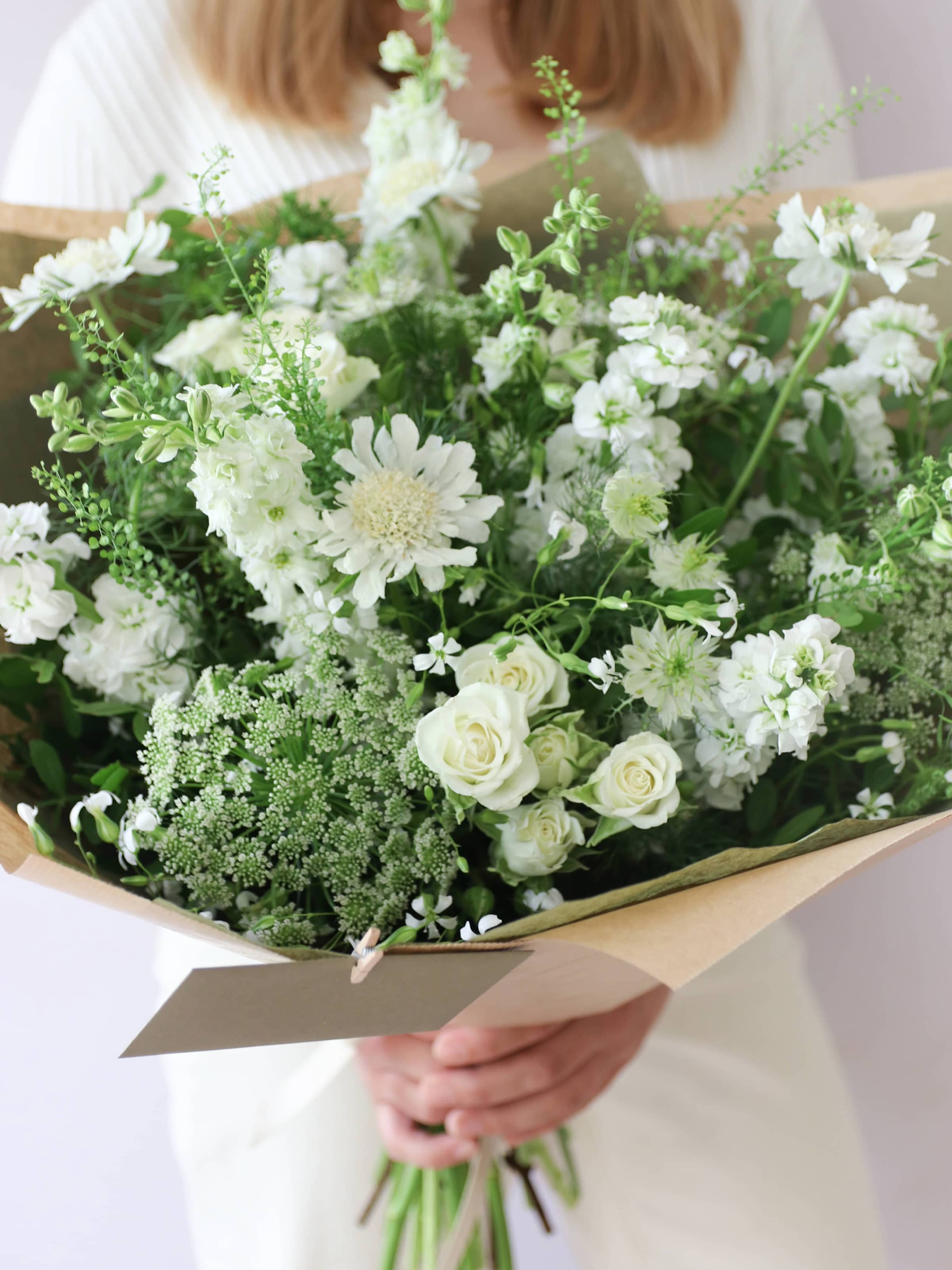 A woman wearing a white jumper and cream jeans, holding a white and green gift-wrapped bouquet of flowers including Delphiniums, Stocks, Roses and Ammi in both hands against a neutral background. The bouquet has a message card and enveloped attached to it with a wooden peg.