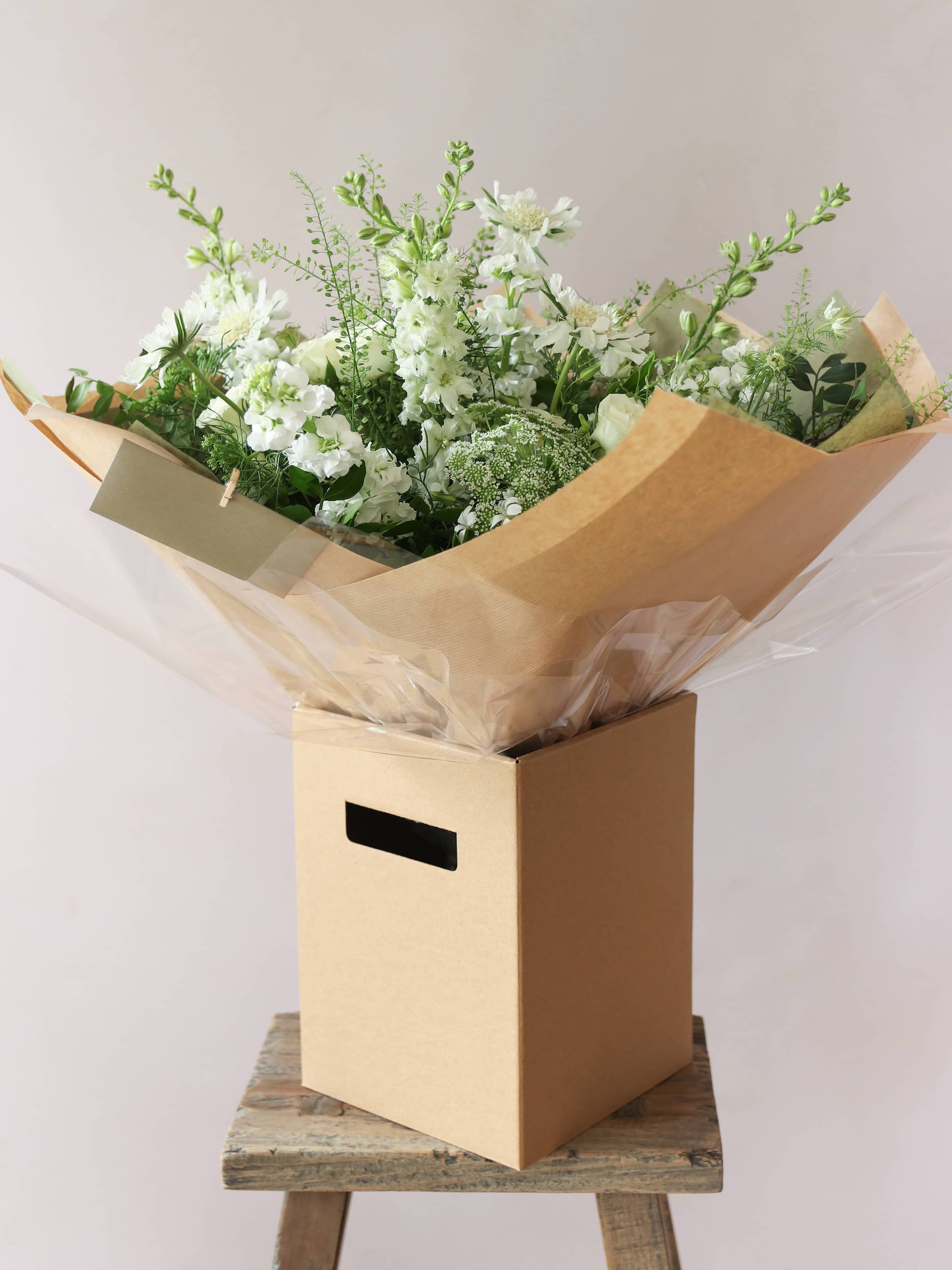 A gift-wrapped bouquet of green and white flowers standing in a kraft brown gift box. The gift box is sitting on a small wooden stool against a neutral background.