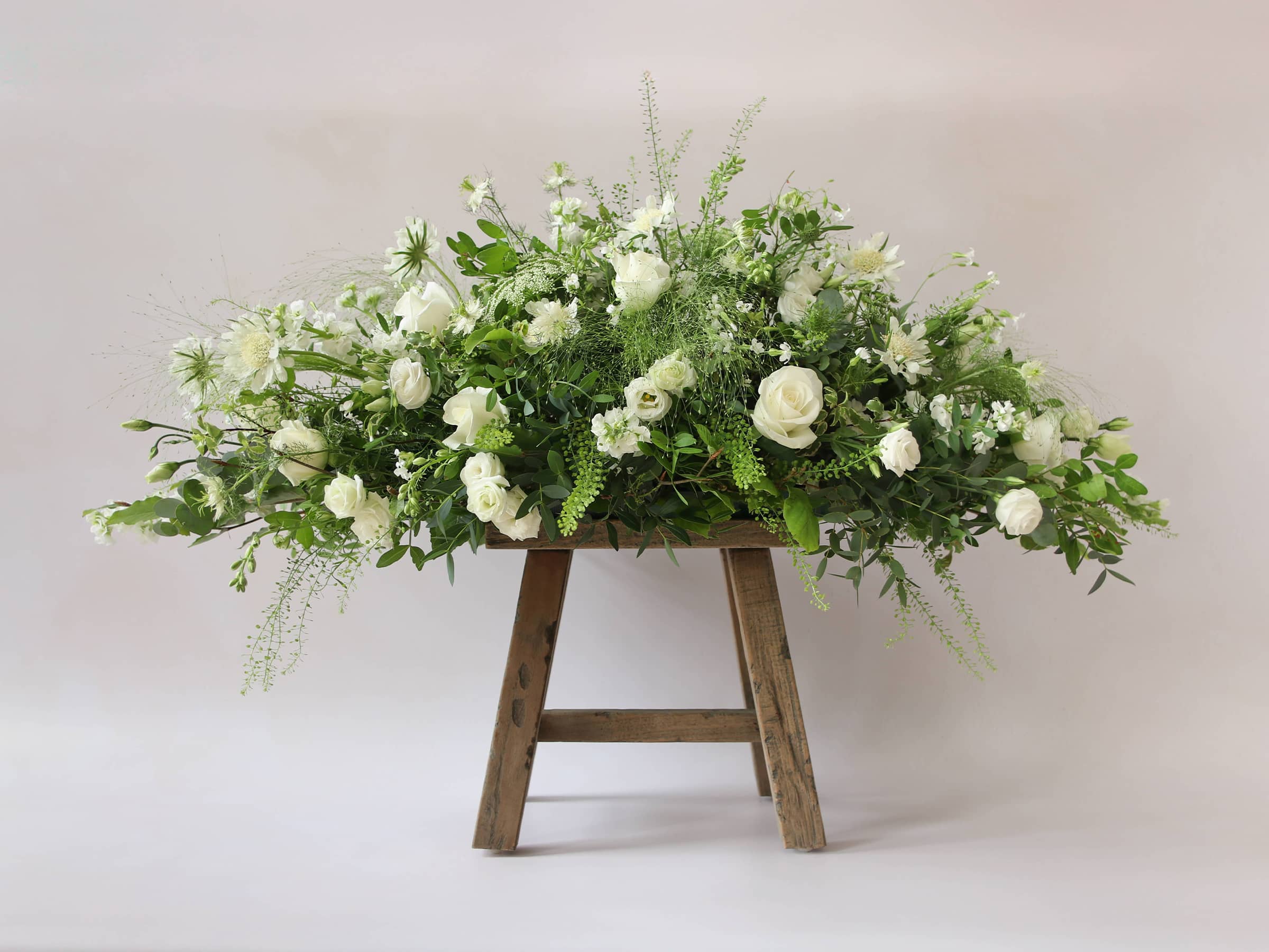 A 3ft coffin spray featuring white and green flowers and foliage. The funeral arrangement is sitting on a rustic wooden stool against a neutral background and features white Roses, Ammi, Stocks and Lisianthus.
