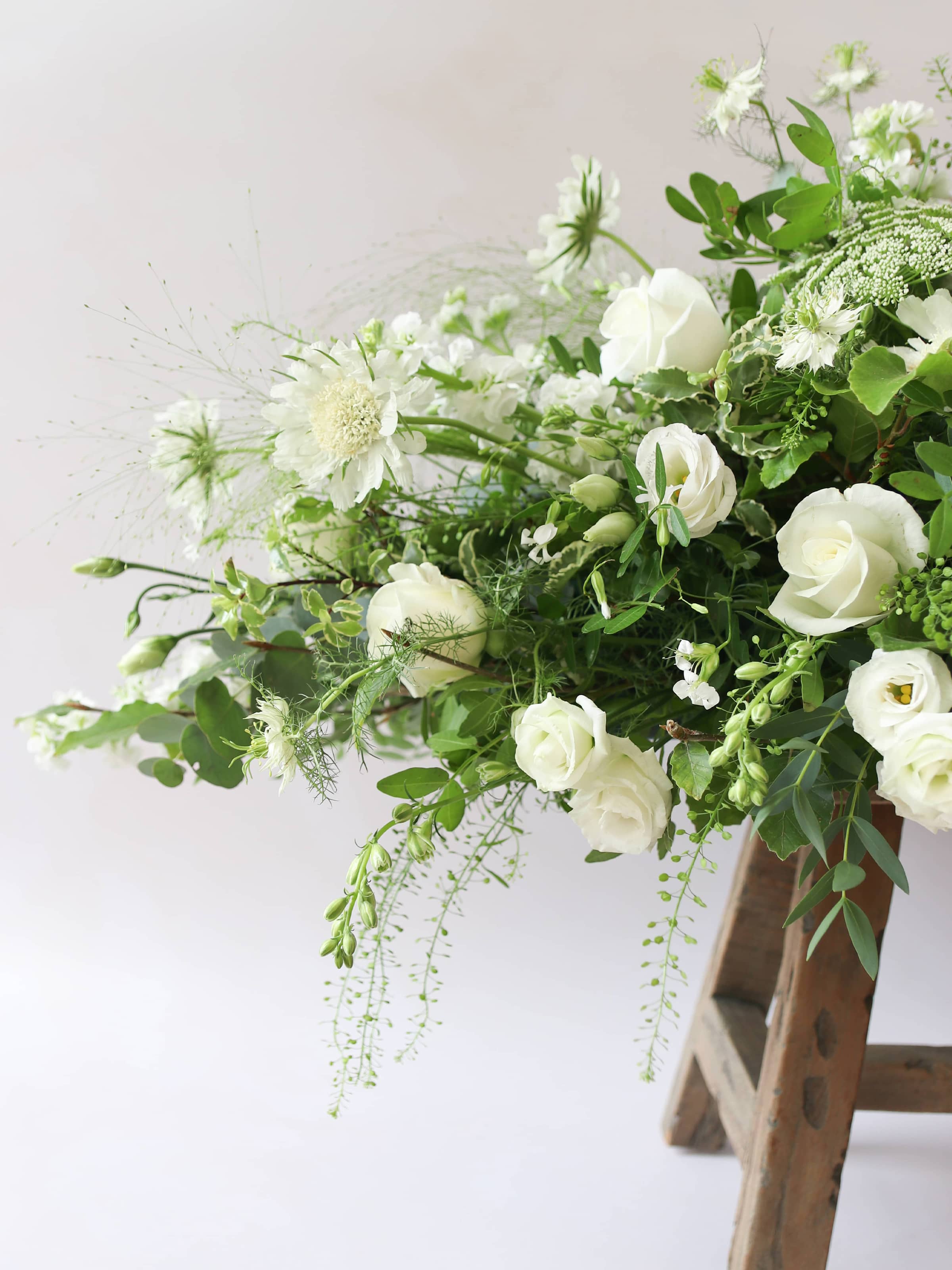 The pointed end of a 3ft coffin spray featuring white and green flowers and foliage. The funeral arrangement is sitting on a rustic wooden stool against a neutral background.