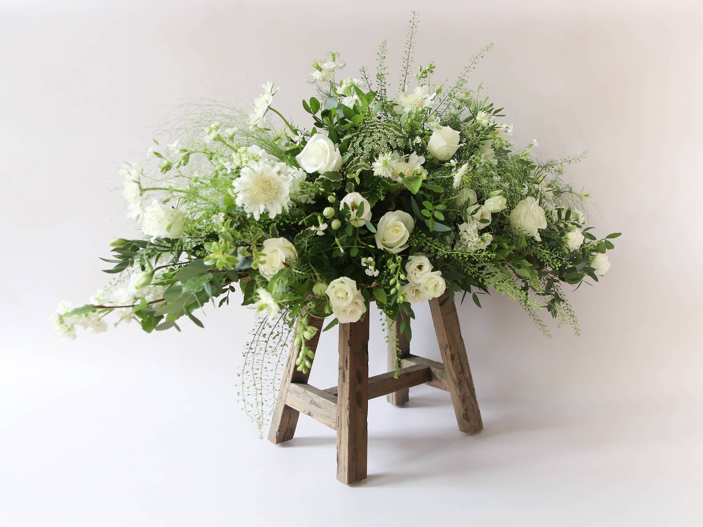 A 3ft coffin spray featuring white and green flowers and foliage. The funeral arrangement is sitting on a rustic wooden stool against a neutral background.