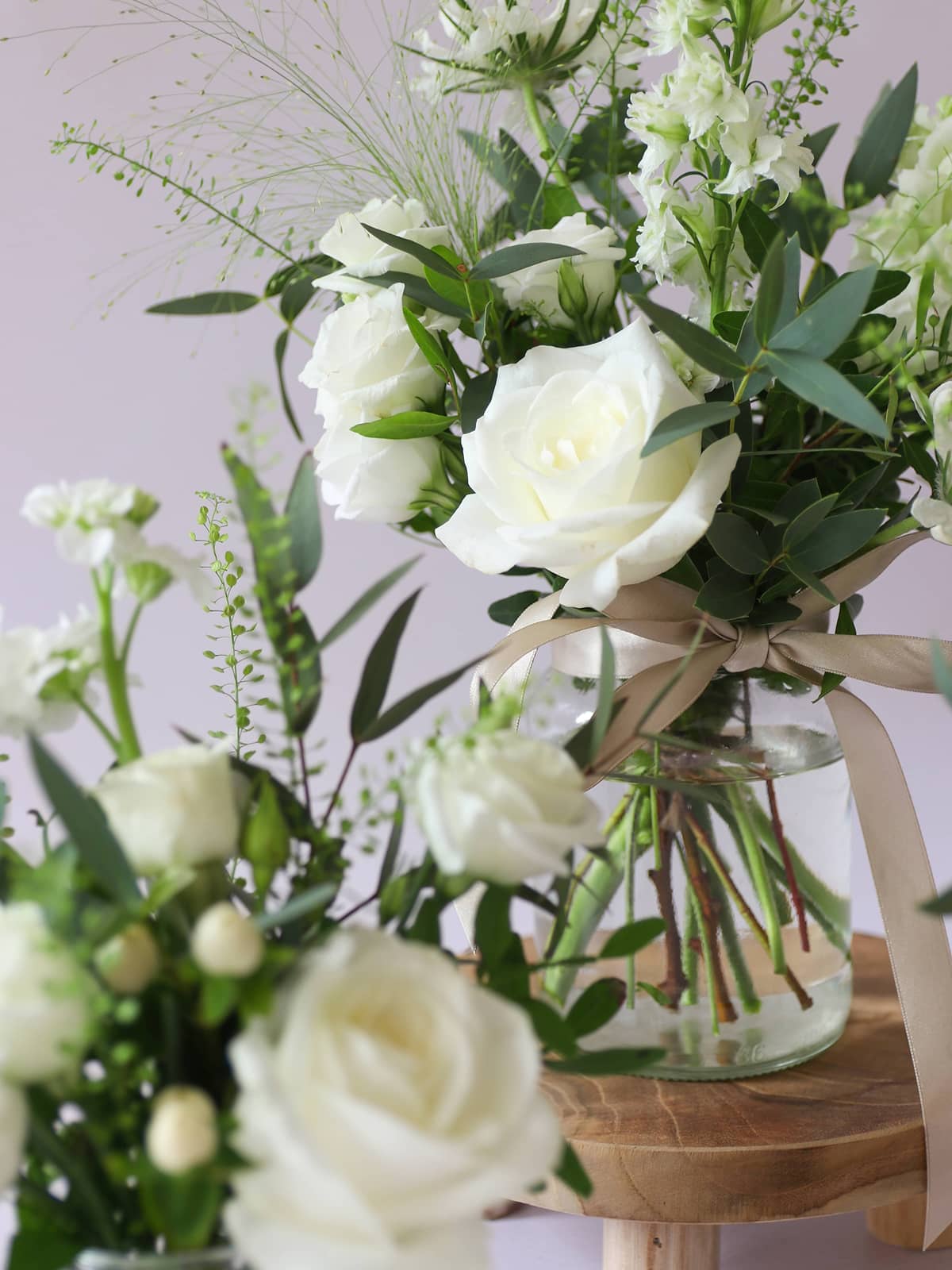 Small bouquet of white roses and greenery in a clear jam jar on a wooden surface with a light purple background.