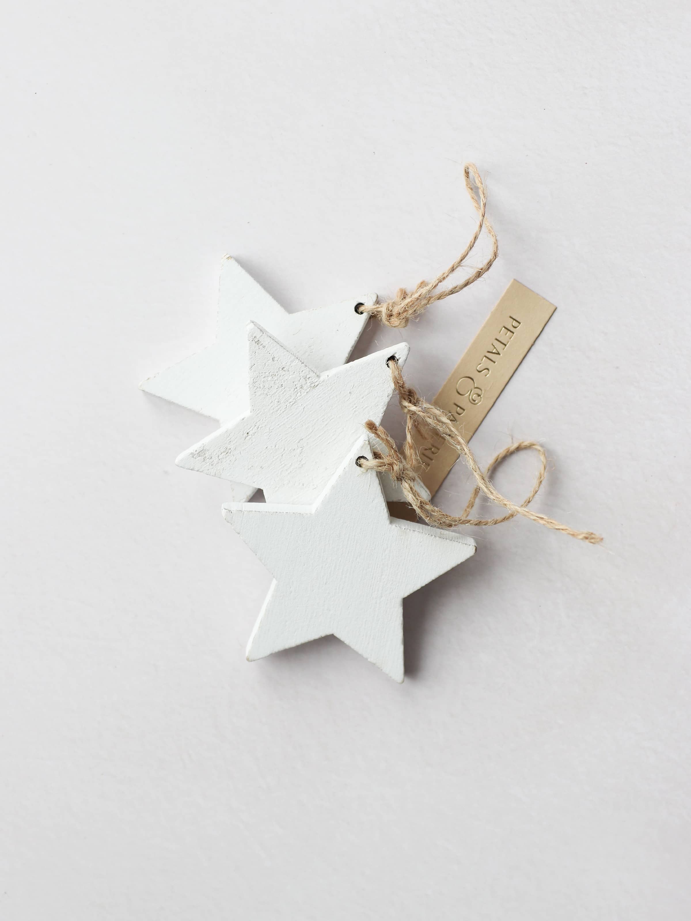 3 white star-shaped wooden ornaments for Christmas wreath making with brown twine tie loops, on a light grey background. The stars are tied together with a Petals & Paperie gift tag.