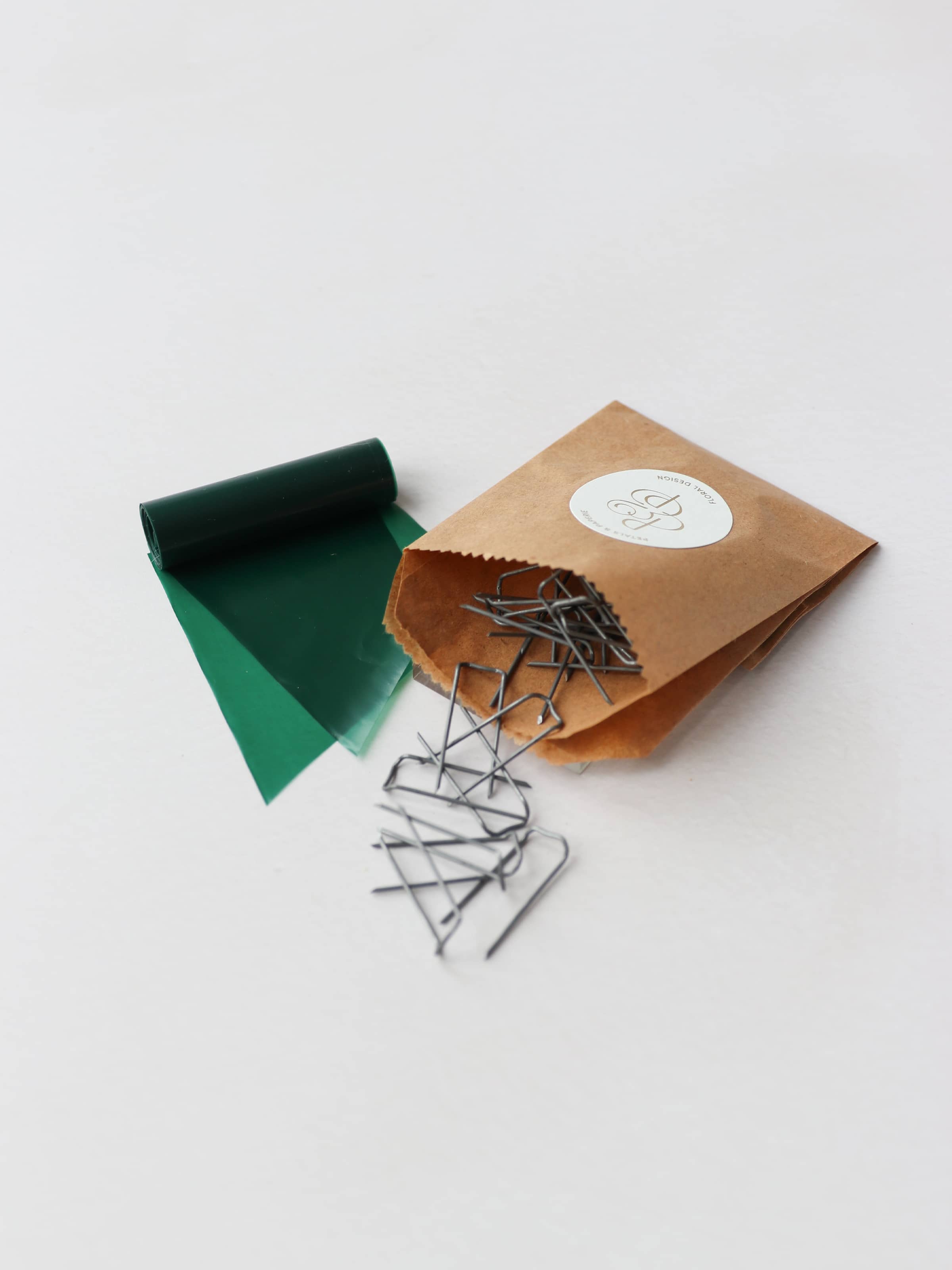 A close up of a kraft paper bag containing metal mossing pins and roll of green wreath wrap, sitting on a neutral background.