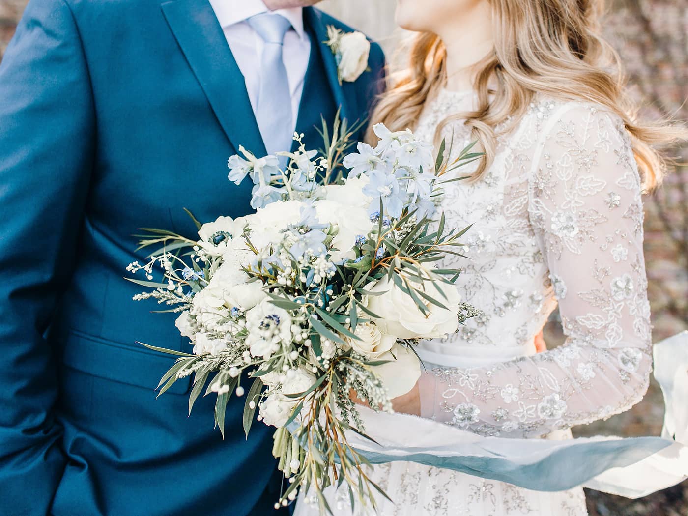 A groom in a blue suit and woman in an embellished white bridal dress holding a bouquet of white and blue flowers with trailing blue ribbons at Middleton Lodge, The Fig House.