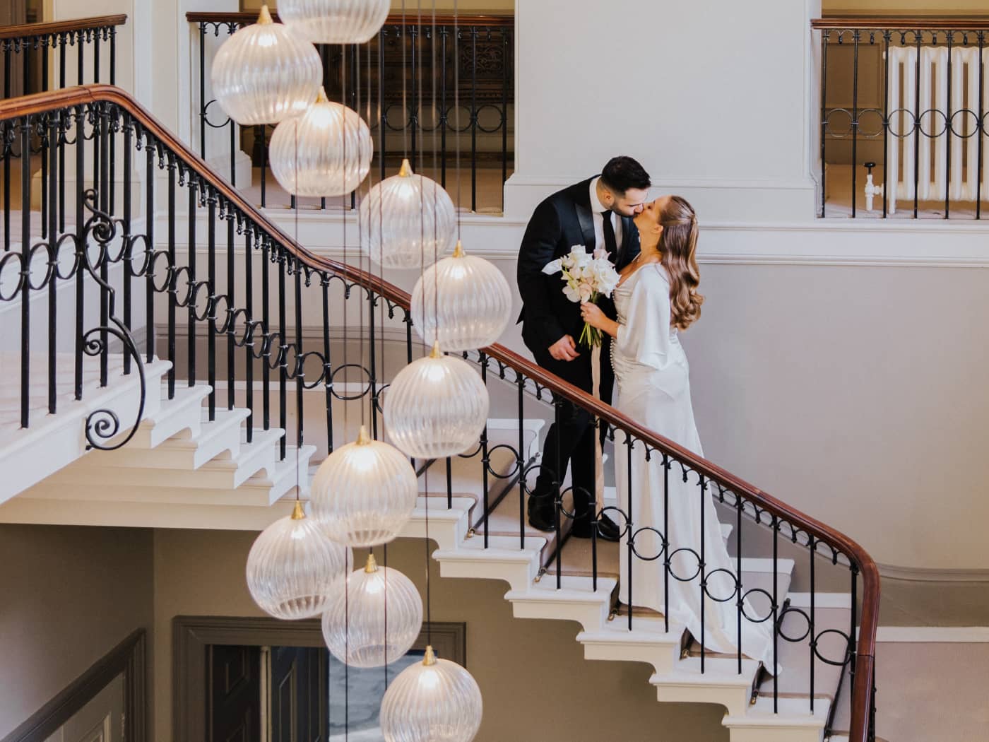 A bride wearing a silk wedding dress carrying a white and blush colour bridal bouquet and a groom in a black suit standing on the staircase at Saltmarshe Hall with a hanging chandelier.