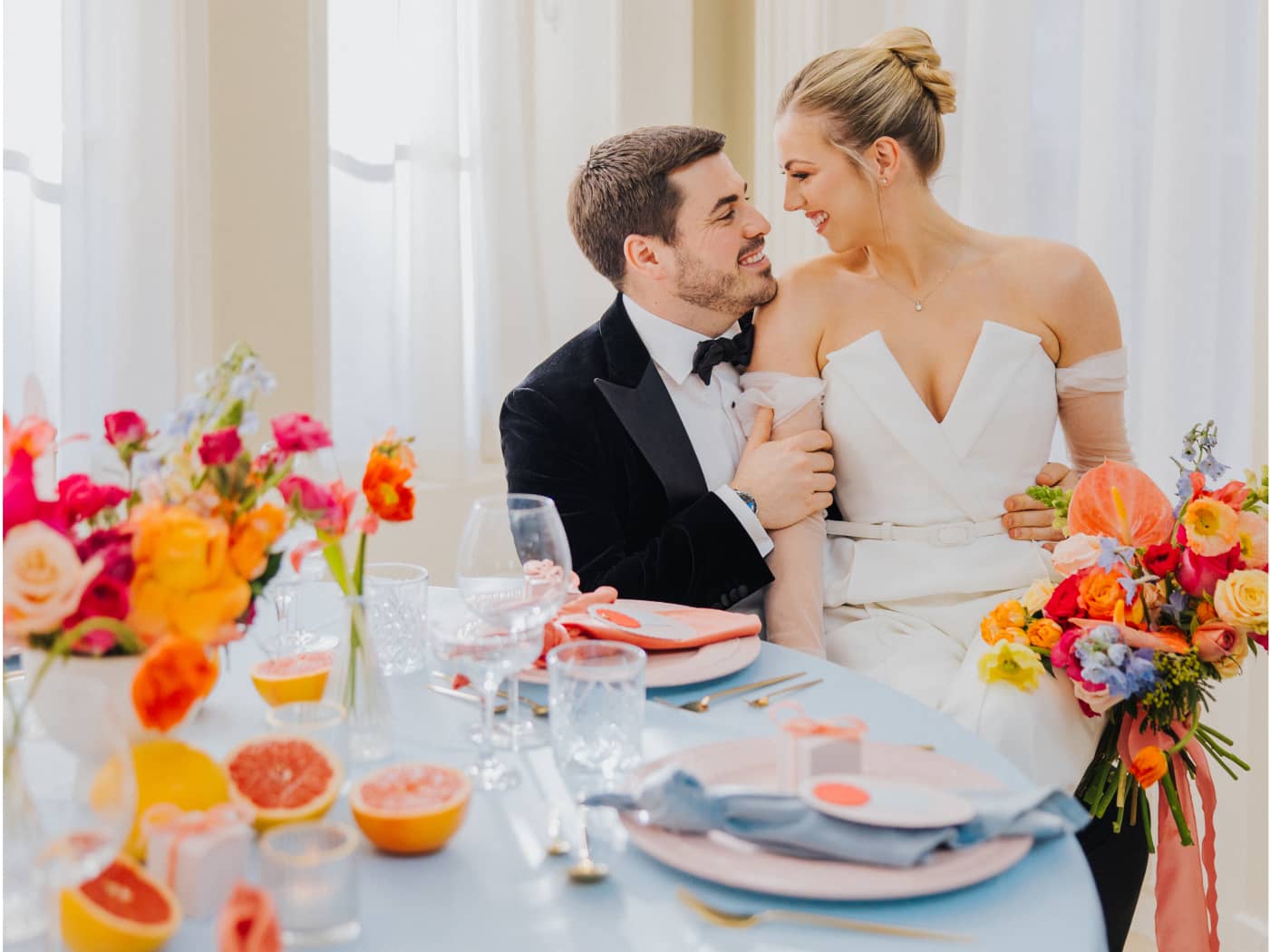 A bride dressed in a white jumpsuit and a groom in a black tuxedo sitting at a round wedding table at Saltmarshe Hall, embracing each other. The bride is carrying a bright and colourful wedding bouquet.