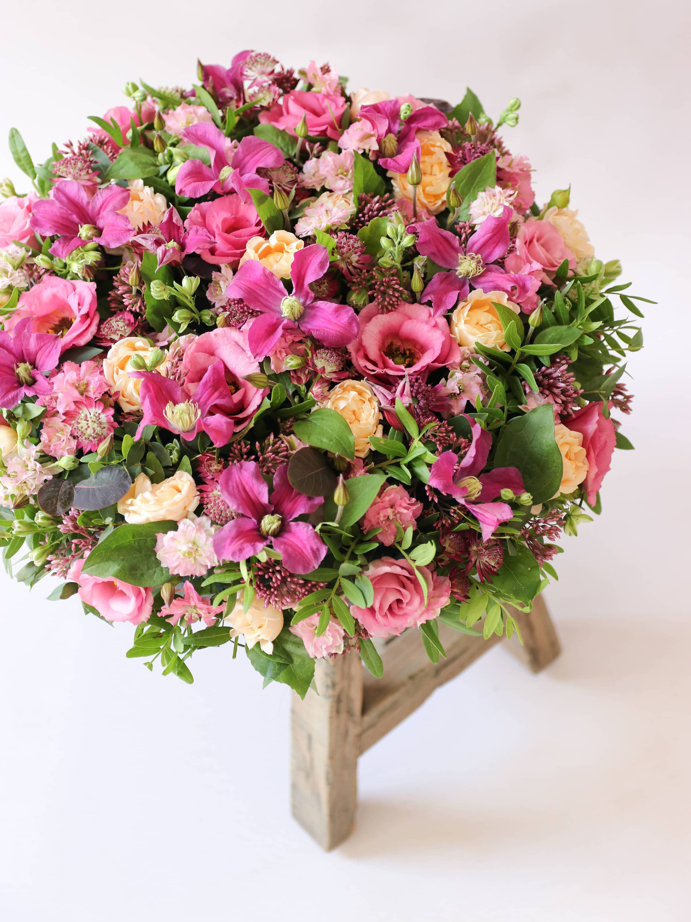 Angled top down side view of a round funeral posy of bright pink and peach flowers with green foliage sitting atop a wooden stool on a white background