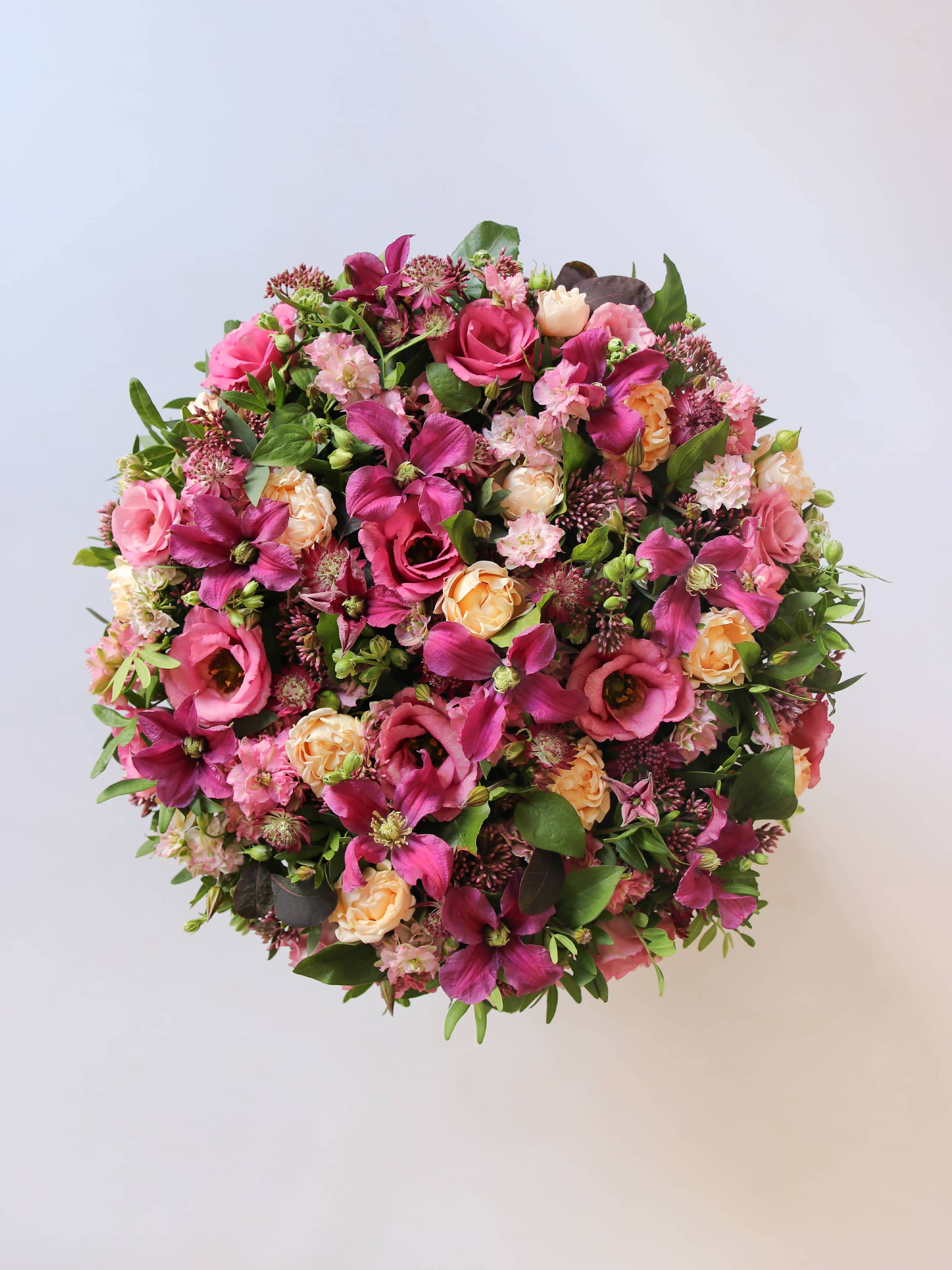 Top down view of a round funeral posy of bright pink and peach flowers with green foliage on a white background