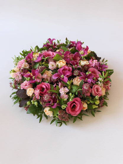 Angled view of a round funeral posy of bright pink and peach flowers with green foliage on a white background