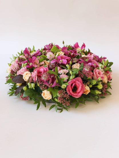 Side view of a round funeral posy of bright pink and peach flowers with green foliage on a white background