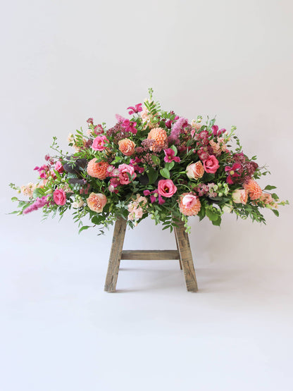 Side view of a double ended coffin spray with bright pink and peach flowers and green foliage, sitting a top a wooden stool on a white background.