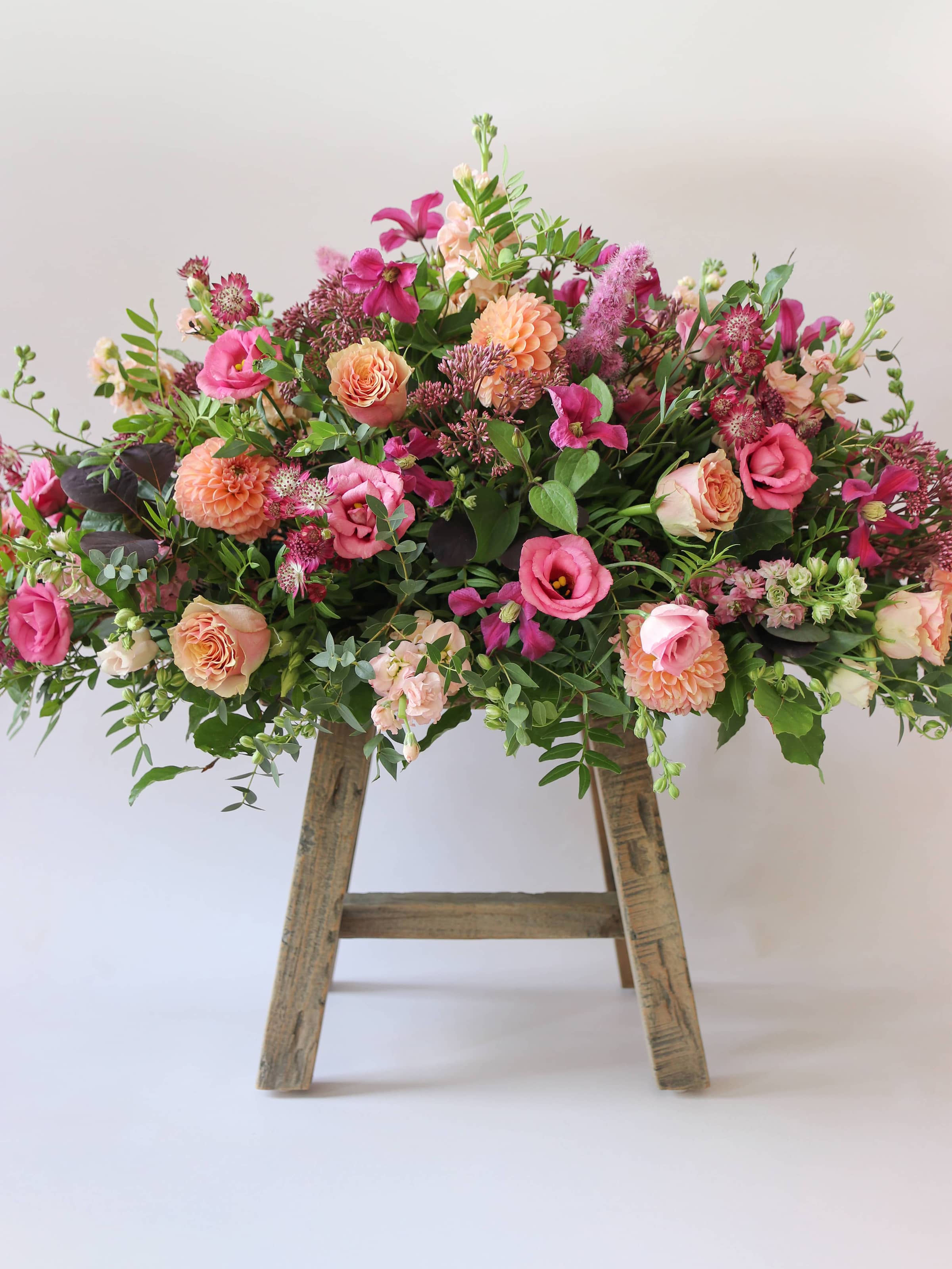 Double ended coffin spray of pink, orange, and green flowers on a wooden stool against a white background