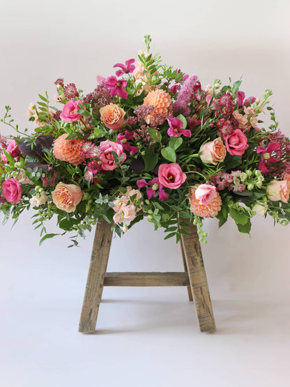 Double ended coffin spray of pink, orange, and green flowers on a wooden stool against a white background