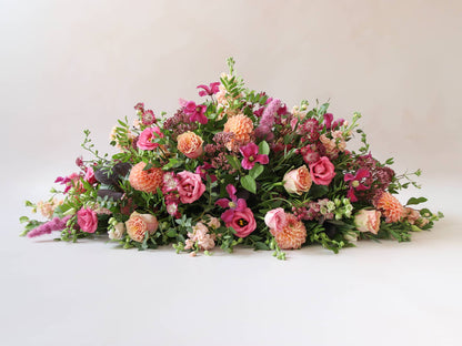 Side view of a double ended coffin spray with bright pink and peach flowers and green foliage, laying on a white background.