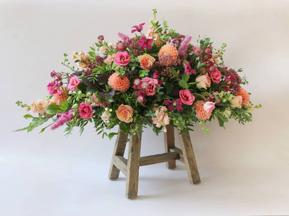 Double ended coffin spray with bright pink and peach flowers and green foliage, viewed at an angle on a wooden stool.