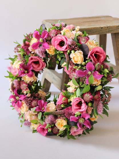 Floral wreath with pink, yellow, and green flowers on a white background with a wooden stool in the background.