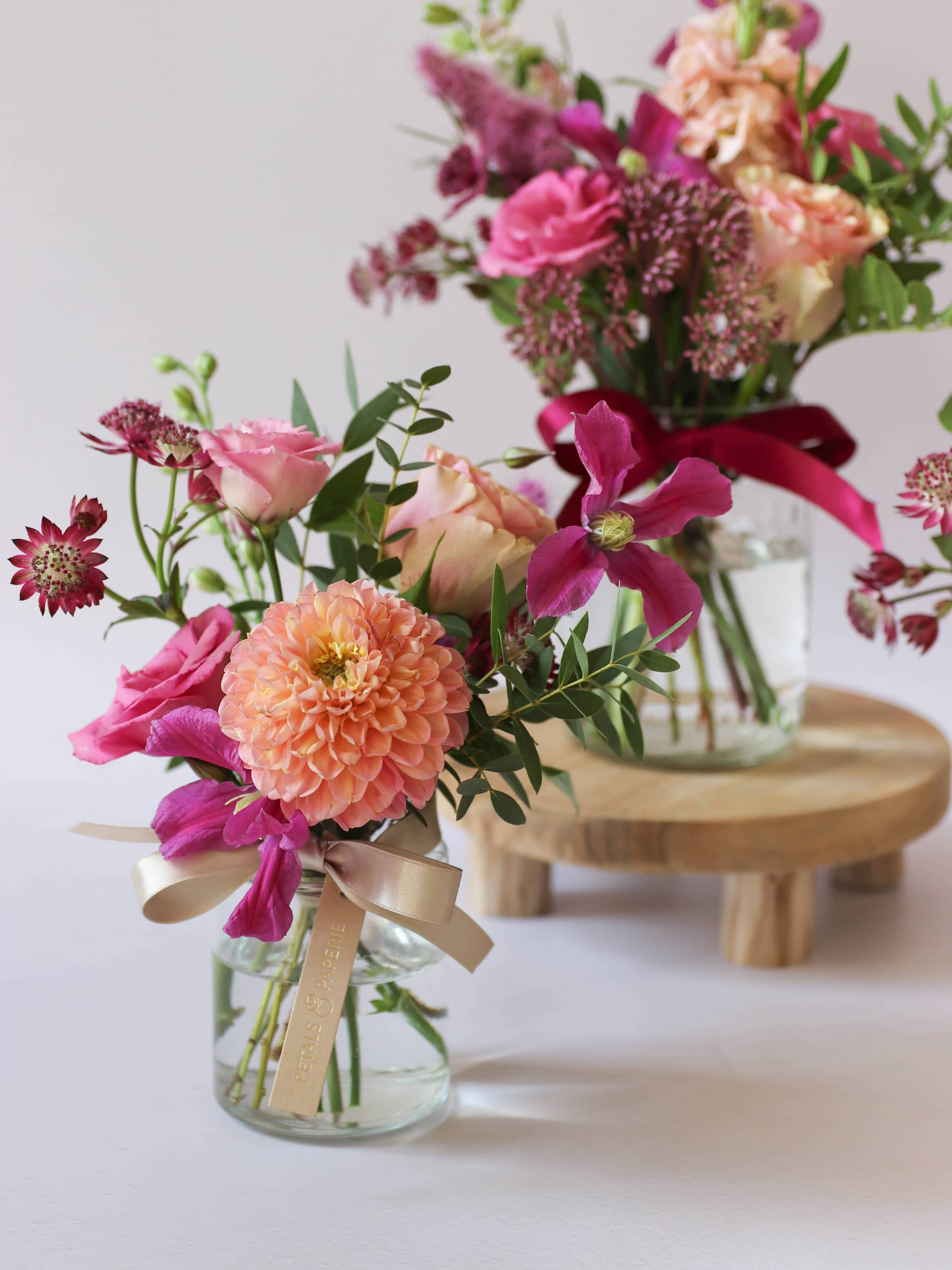 Two bright pink and peach floral arrangements in glass jars with ribbons on a white background