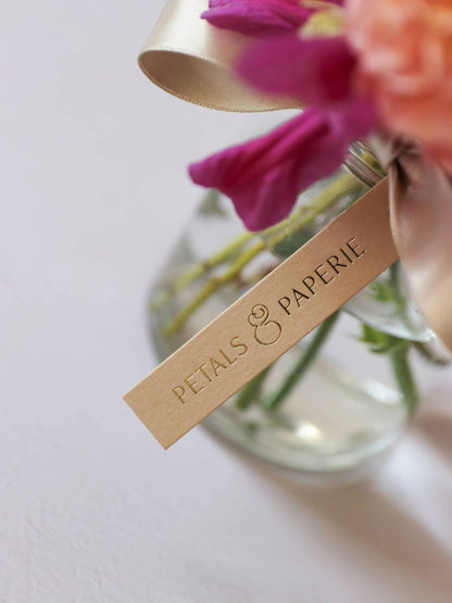Glass vase with flowers and a 'Petals & Paperie' tag on a light background