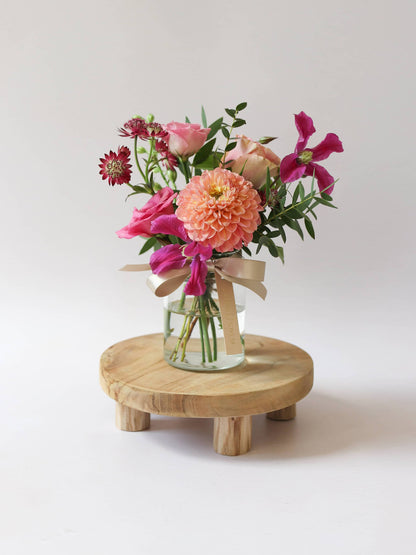 A single jam jar arrangement of bright pink and peach flowers, with a silk ribbon on top of a small wooden stool