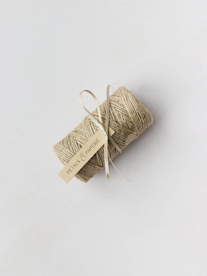 A spool of brown kraft mossing twine tied with a cream satin ribbon and a Petals & Paperie gift tag, sitting on a neutral background.
