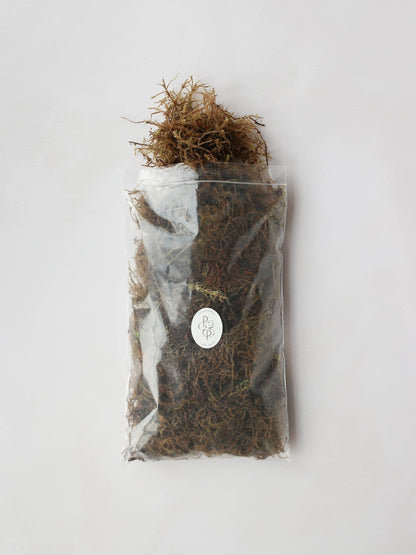 A bag of sphagnum moss packaged in an air-tight plastic resealable bag. The bag has a branded Petals & Paperie sticker on the front and the moss is trailing out of the top of the open bag.