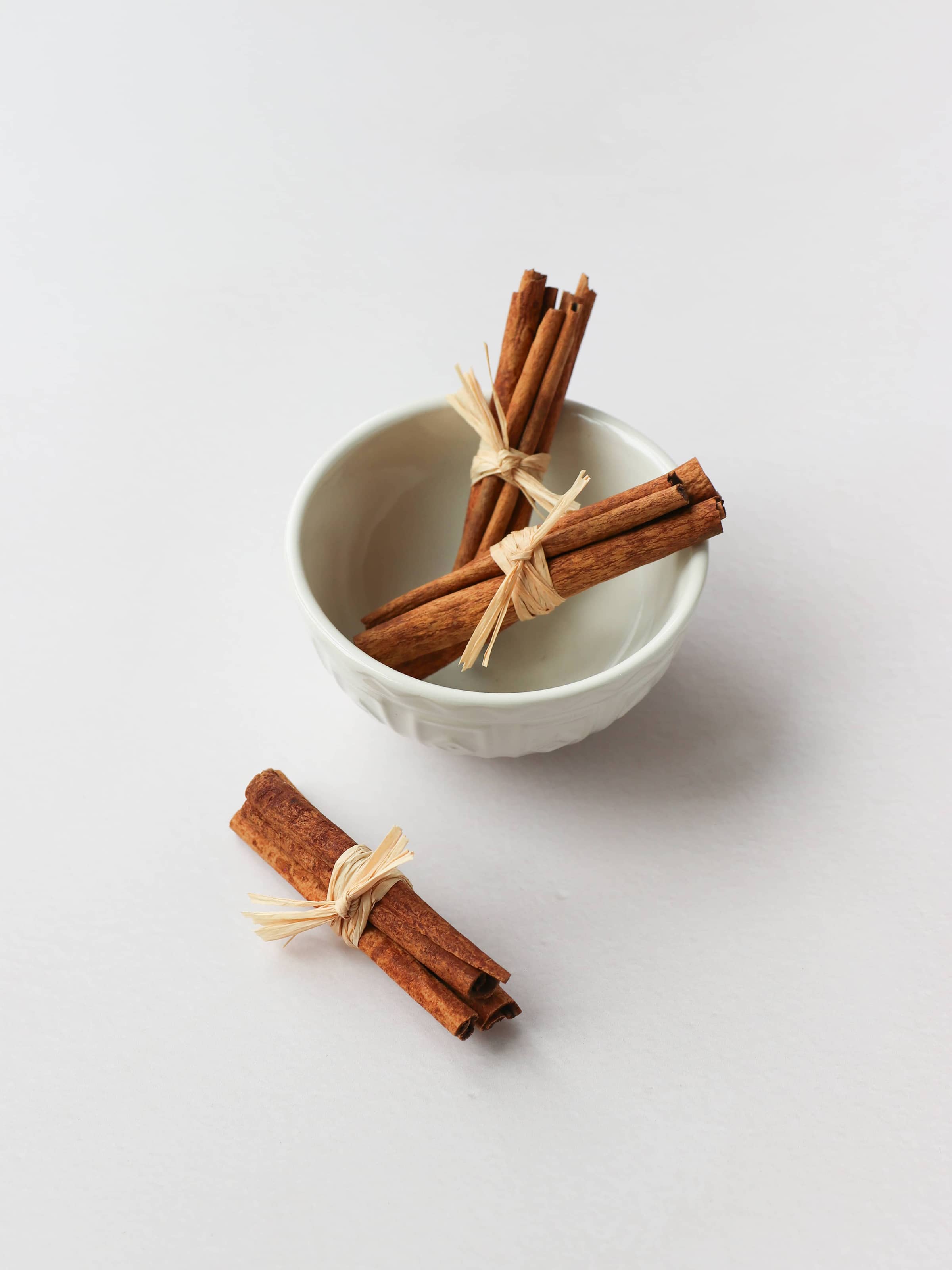 Three bundles of cinnamon sticks tied with raffia for Christmas wreath making, in a decorative cream bowl on a neutral background.