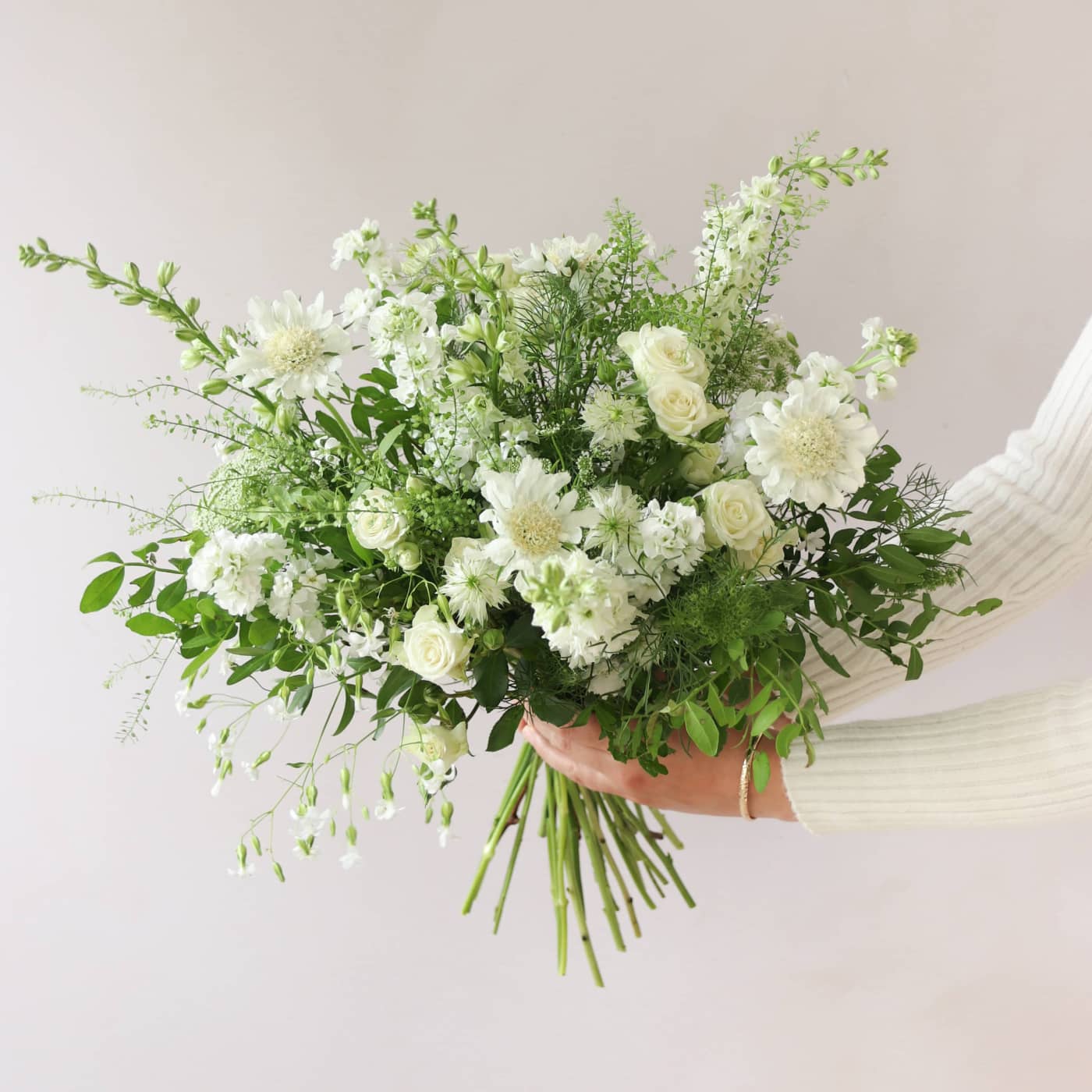 A bouquet of white and green flowers held out to the side by a person against a neutral background.