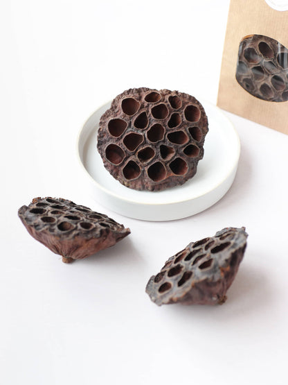 Three dried brown lotus seed pods for Christmas wreath making, styled around a white decorative bowl on a neutral background. There is a brown kraft bag of lotus seed pods in the background.