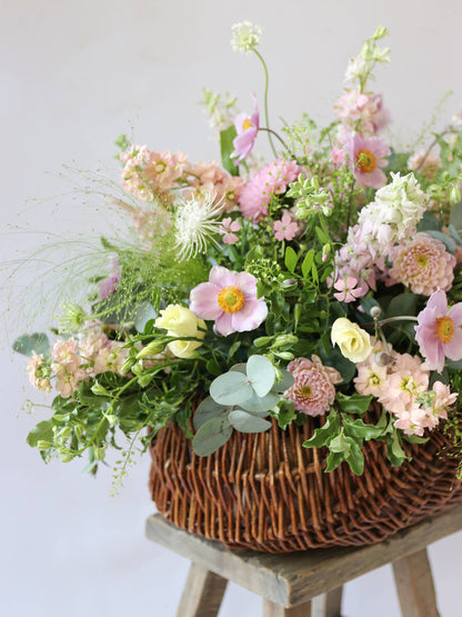 A side view of a wicker basket arrangement featuring pastel pink and yellow flowers. The basket arrangement is placed on a rustic wooden stool against a neutral background and features pink Dahlias, yellow Lisianthus and peach Stocks.