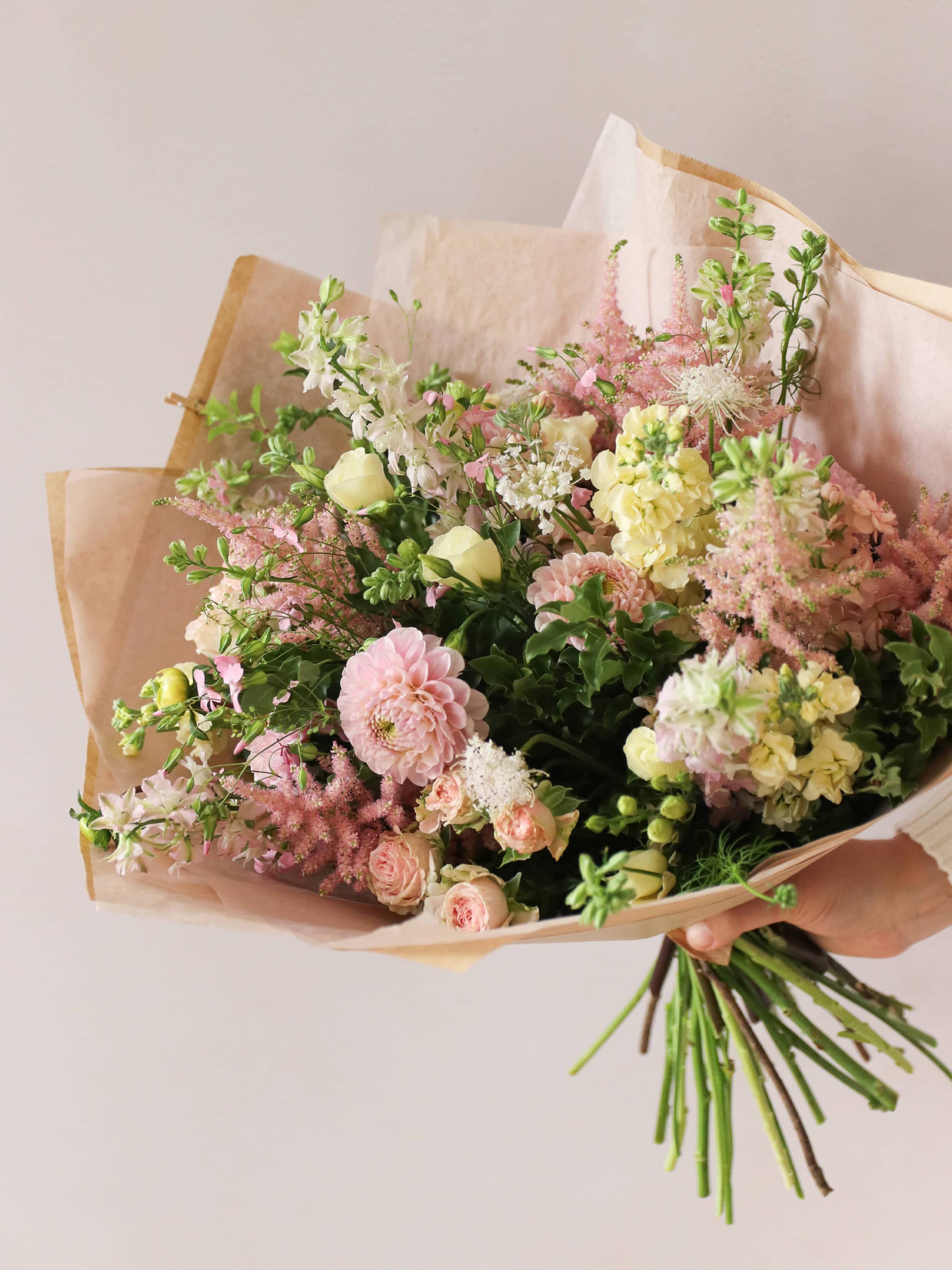 A person holding a pastel yellow and pink gift-wrapped bouquet of flowers including Astilbe, Dahlias, Roses and Lisianthus in one hand against a neutral background.