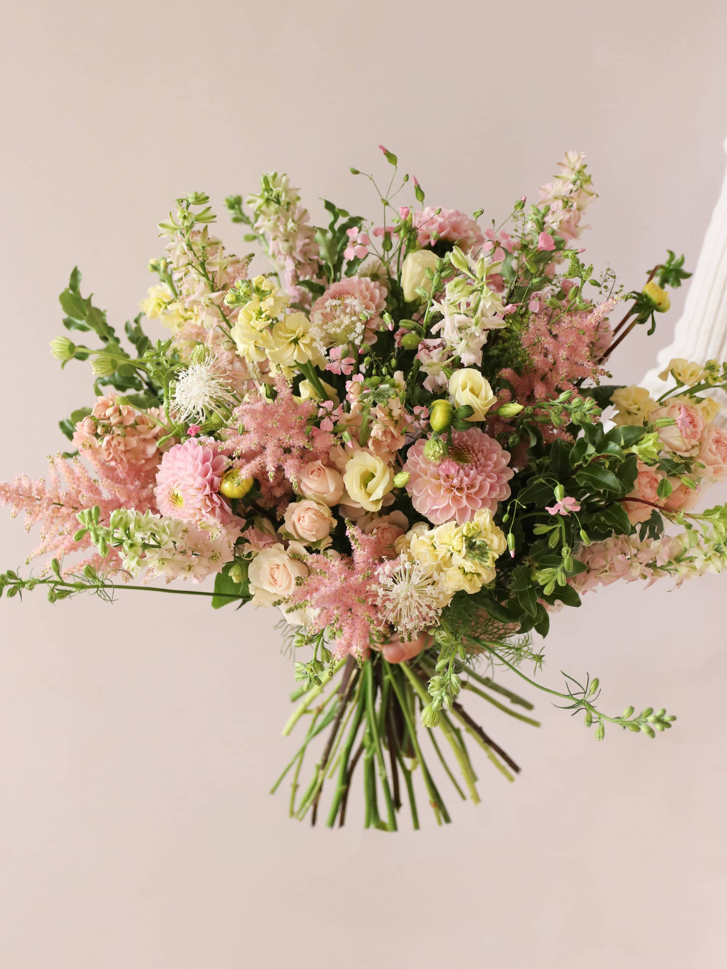 A person holding a pastel yellow and pink bouquet of flowers including Astilbe, Dahlias, Roses and Lisianthus in both hands against a neutral background.