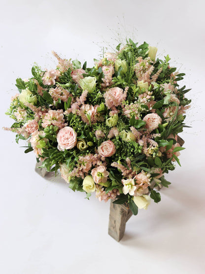 Heart shaped funeral posy with pink and green colours on a white background sitting on a wooden stool