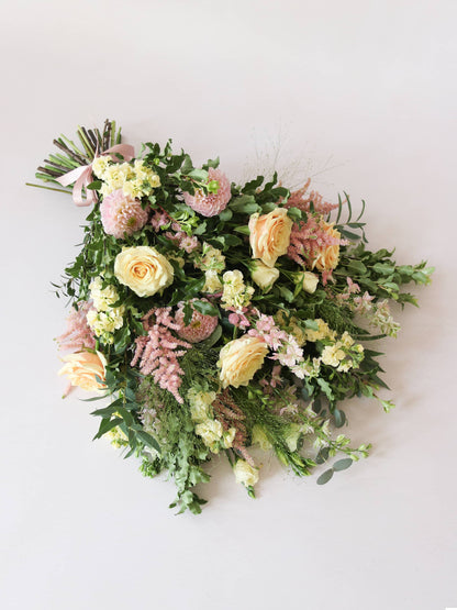 A top down view of a pastel tied funeral sheaf containing pastel pink, peach and yellow flowers, tied with a satin pink ribbon.