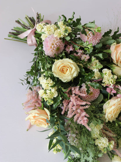 A top down view of a pastel tied funeral sheaf containing pastel pink, peach and yellow flowers, tied with a satin pink ribbon.