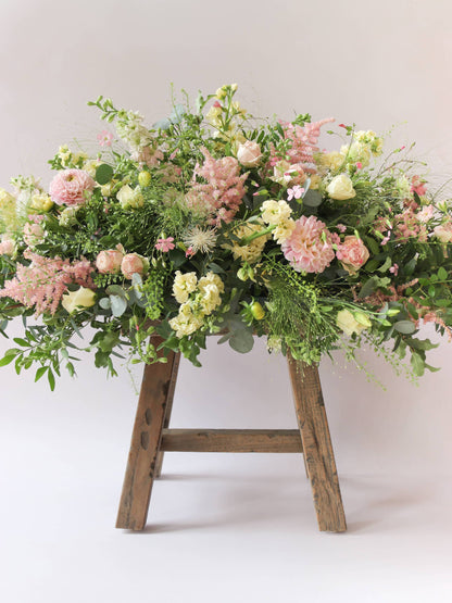 A side view of a 3ft coffin spray featuring pastel pink, yellow and peach flowers and foliage. The funeral arrangement is sitting on a rustic wooden stool against a neutral background and features Roses, Astlibe, Stocks and Lisianthus. 