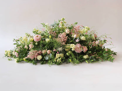 A side view of a 3ft coffin spray featuring pastel pink, yellow and peach flowers and foliage. The funeral arrangement is sitting against a neutral background and features Roses, Astlibe, Stocks and Lisianthus. 